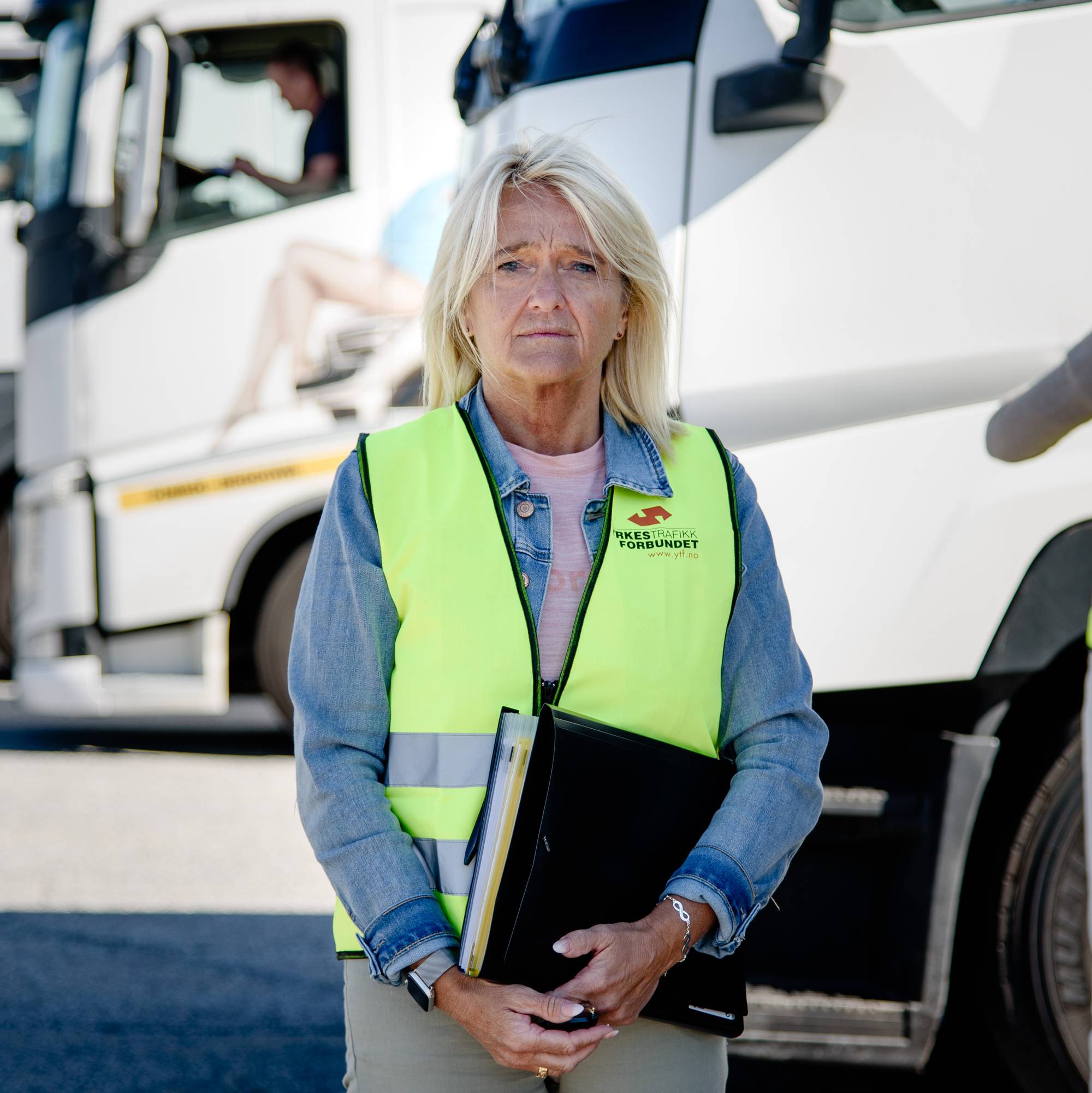 En kvinne med lyst hår, blå genser og en gul refleksvest står foran hvite parkerte lastebiler, Bak henne annen kvinne også med gul refleksvest står med ryggen til ved en lastebil. 