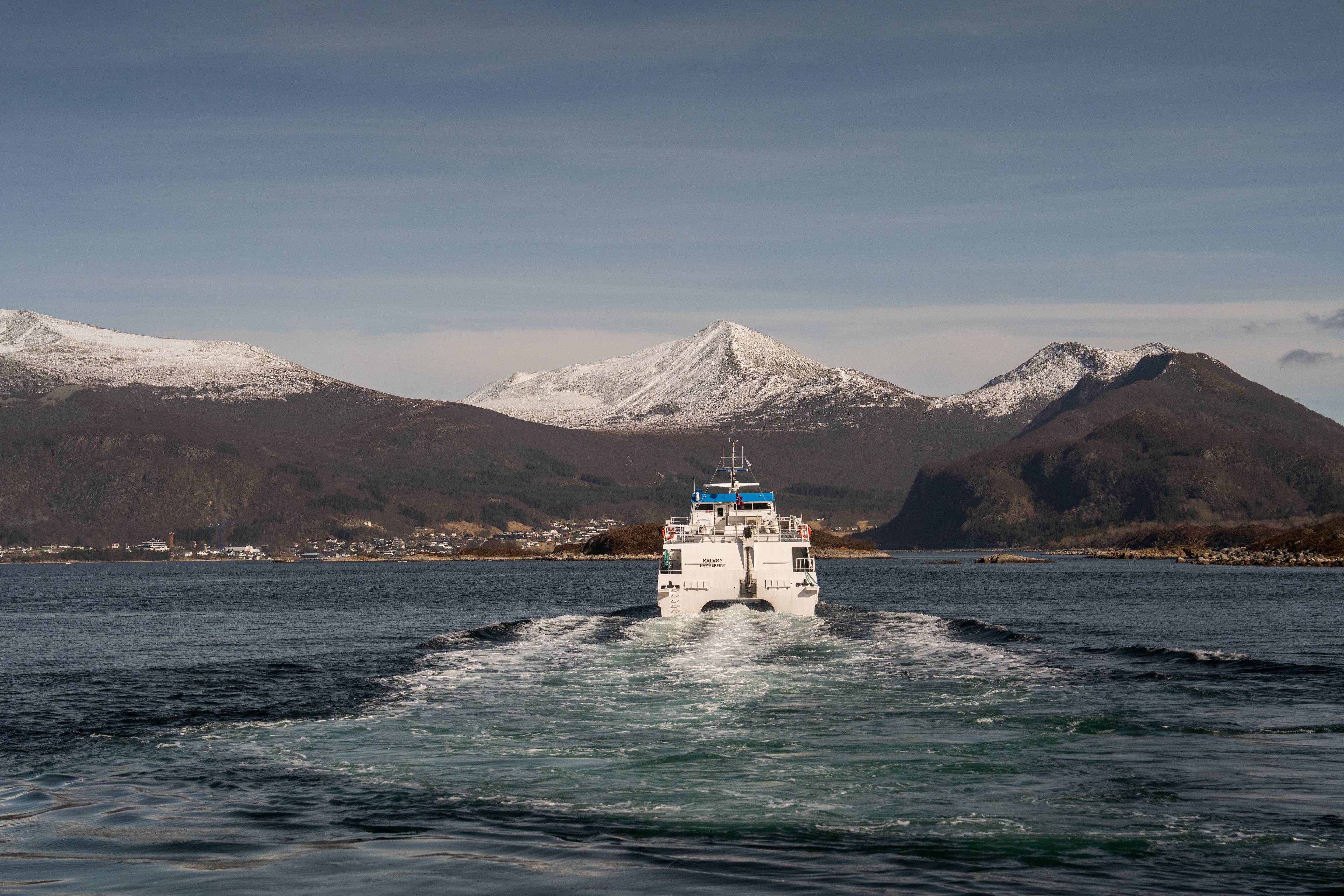 Et hvitt fergeskip seiler bortover en vannflate med snødekte fjell i bakgrunnen. Skipet lager et tydelig kjølvann bak seg. Det er flere andre fartøy synlige i det fjerne, samt en bygd som ligger langs kysten. Himmelen er overveiende klar med noen lette skyer. (Bildebeskrivelsen er laget av en KI-tjeneste)