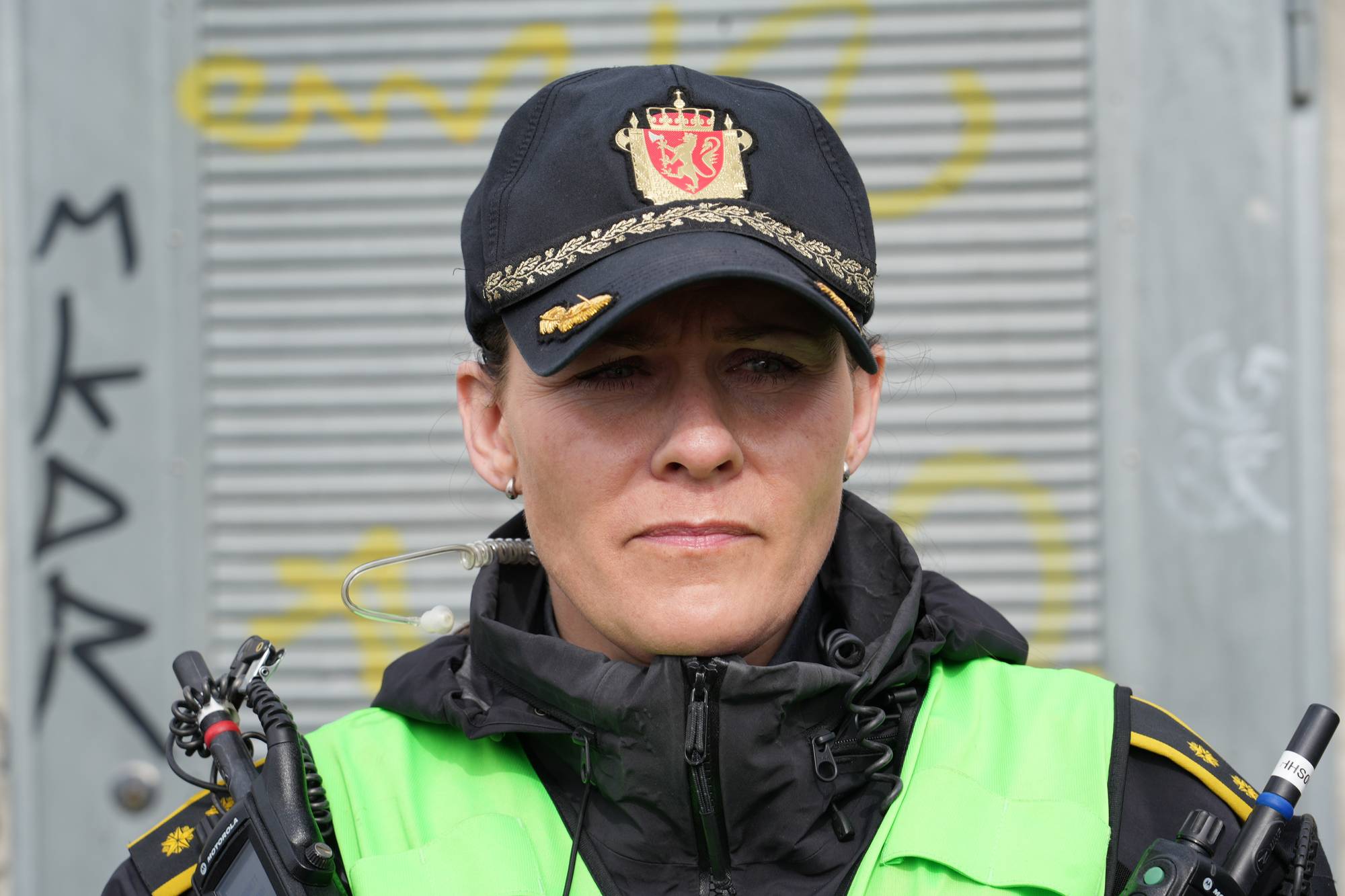 Politibetjent i uniform med caps og refleksvest foran en vegg med graffiti. (Bildebeskrivelsen er laget av en KI-tjeneste)