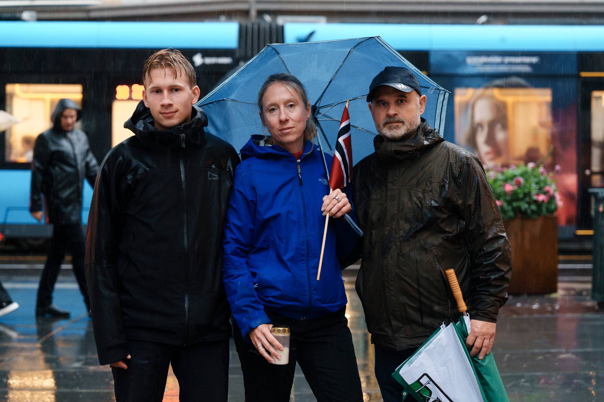 Tre personer står med en paraply og et norsk flagg ved en trikk i regnvær. (Bildebeskrivelsen er laget av en KI-tjeneste)