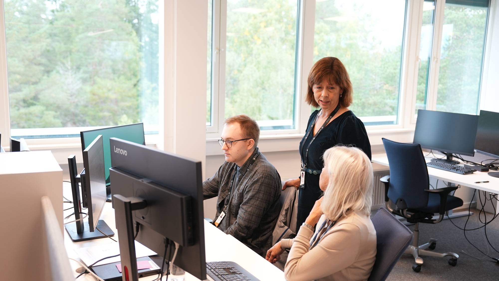 Kvinne i svart kjole snakker med to ansatte ved datamaskiner