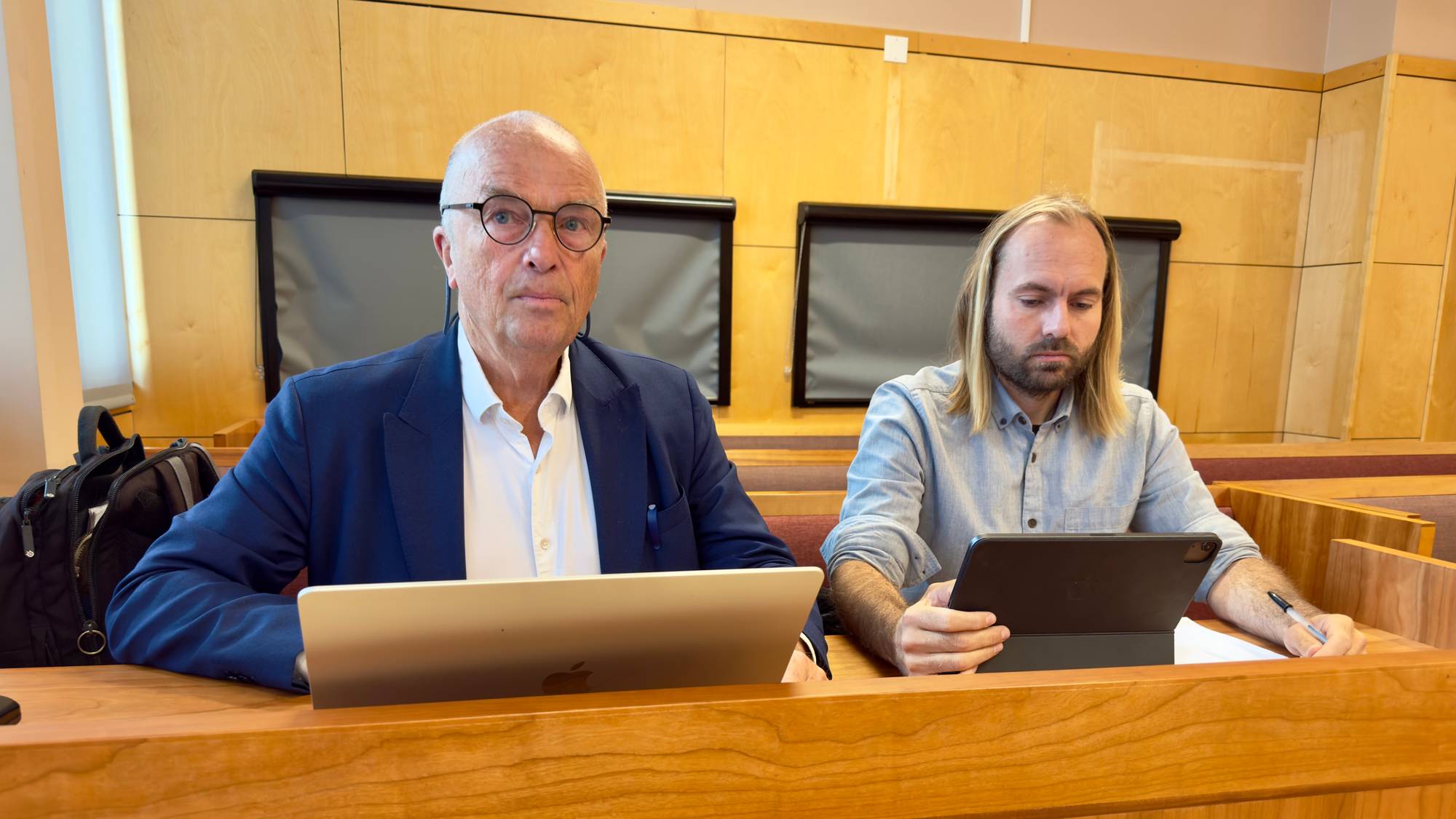 To menn sitter ved en pult i en rettssal. De arbeider på hver sin bærbare enhet. (Bildebeskrivelsen er laget av en KI-tjeneste)