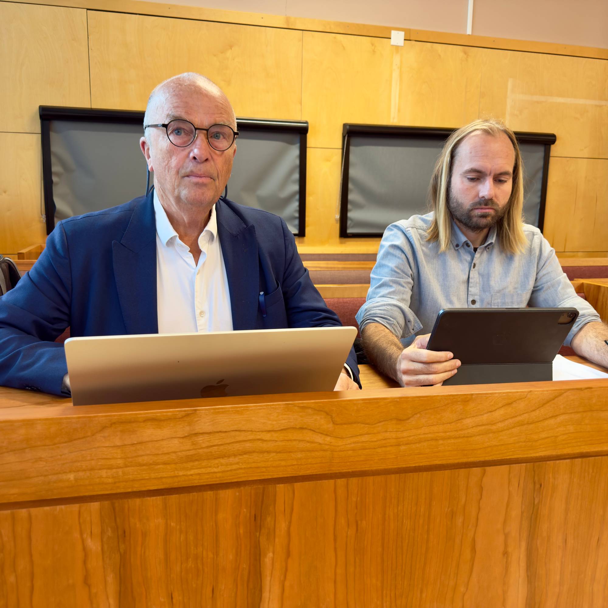 To menn sitter ved en pult i en rettssal. De arbeider på hver sin bærbare enhet. (Bildebeskrivelsen er laget av en KI-tjeneste)