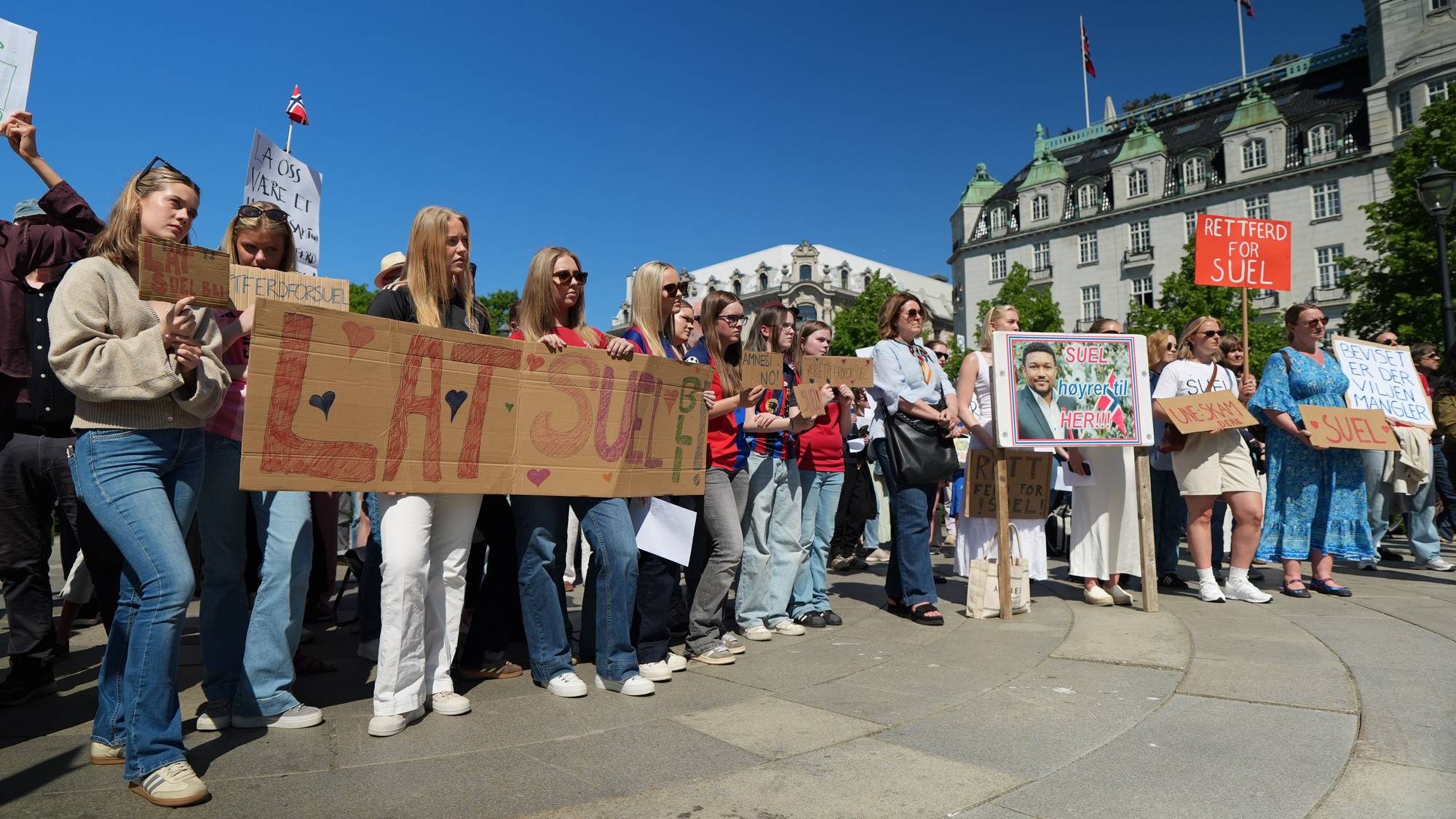 En gruppe demonstranter holder skilt. Et stort skilt i papp med teksten "LAT SUEL BLI" er synlig. En kvinne holder et bilde av en mann. Andre skilt bærer tekster som "Rettferd for Suel".(Bildebeskrivelsen er laget av en KI-tjeneste)
