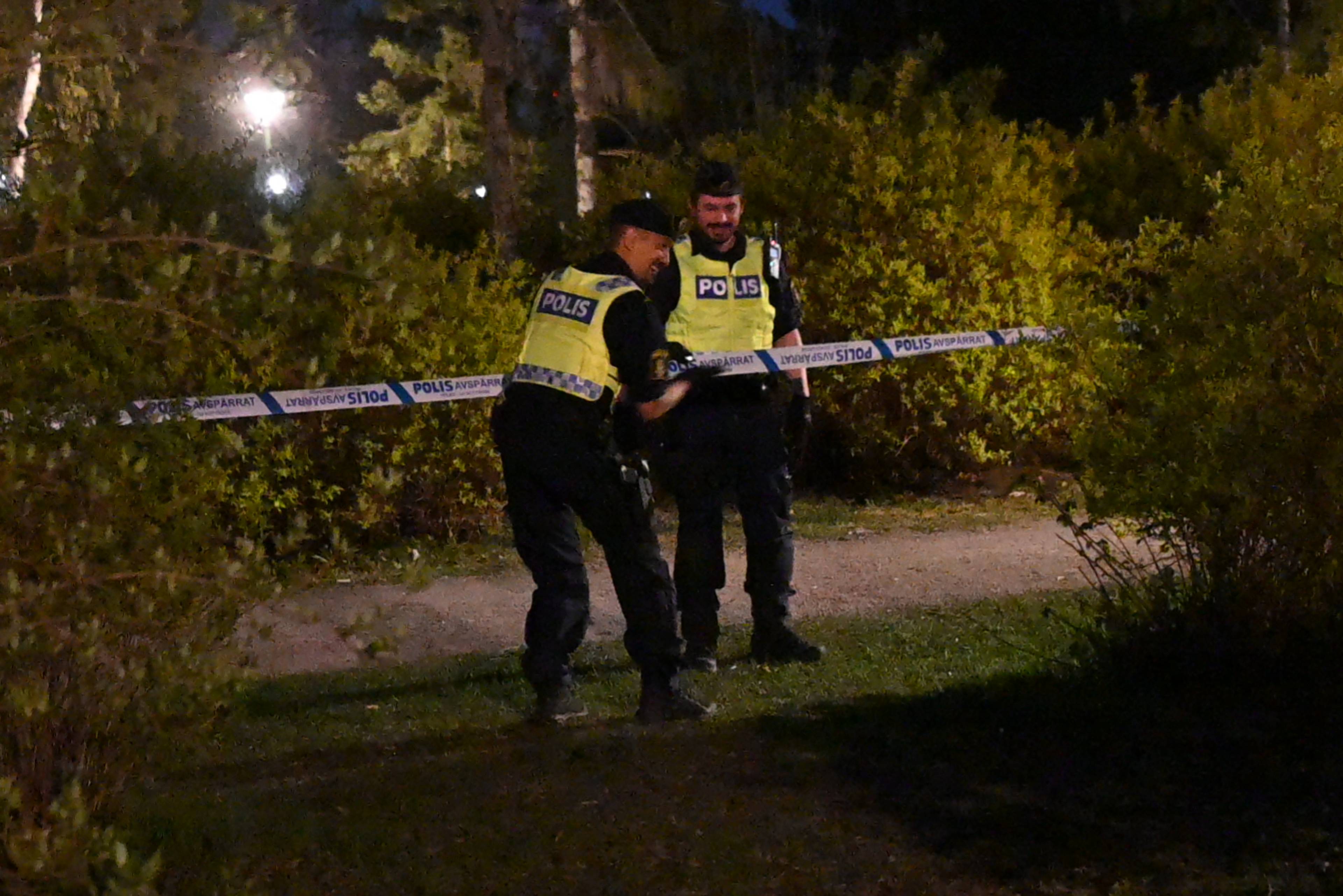 To politibetjenter står ved et avsperringsbånd i en park om kvelden.  De har gule vester med påskriften "POLIS". Bak dem er det buskas og trær. Båndet har teksten "POLIS AVSPRRAT".