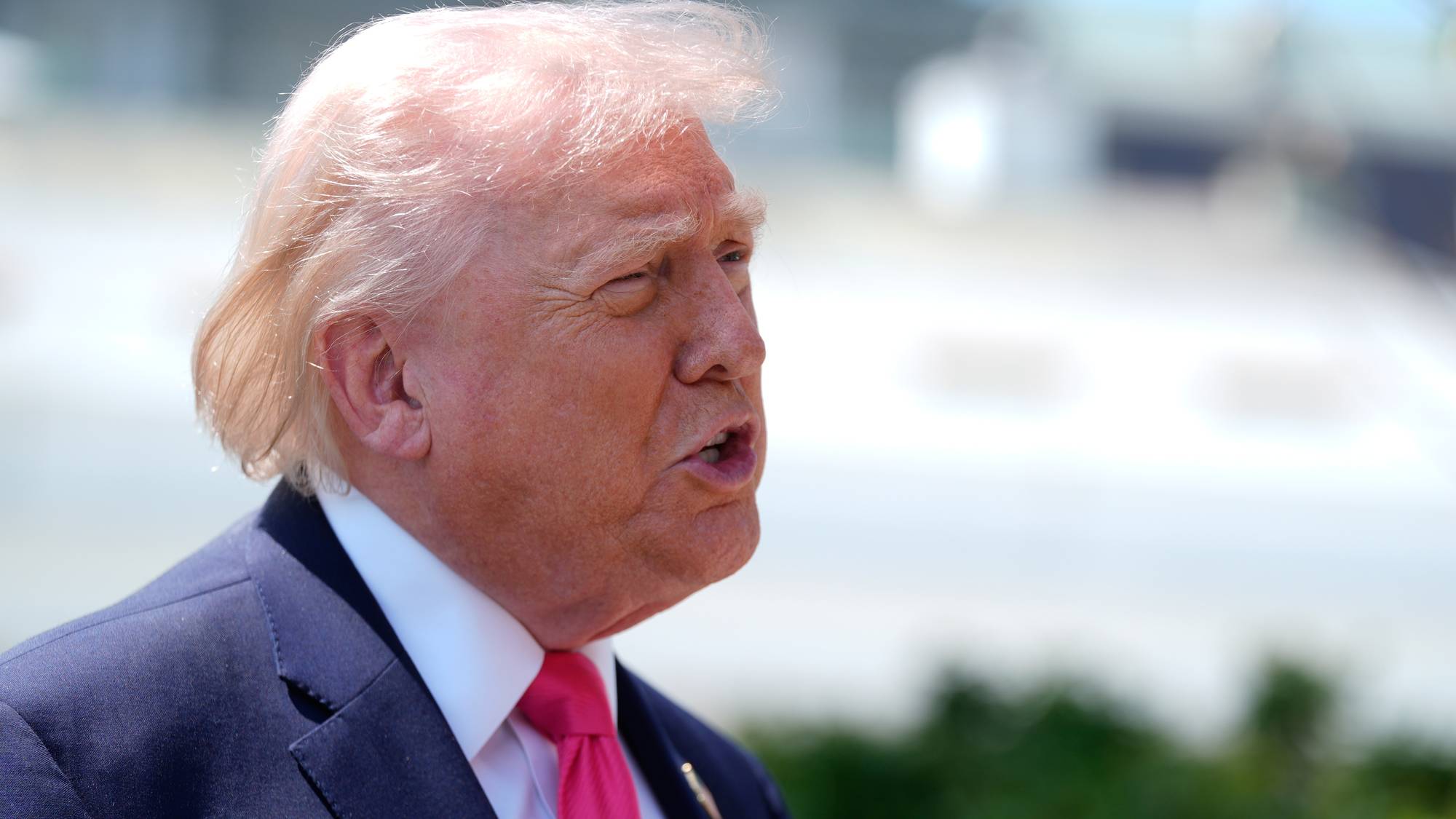 Donald Trump i mørk dress med lys skjorte og en rosa slips. Han ser til høyre og munnen er åpen. I bakgrunnen er det en uskarp hvit bygning og grønne trær. (Bildebeskrivelsen er laget av en KI-tjeneste)