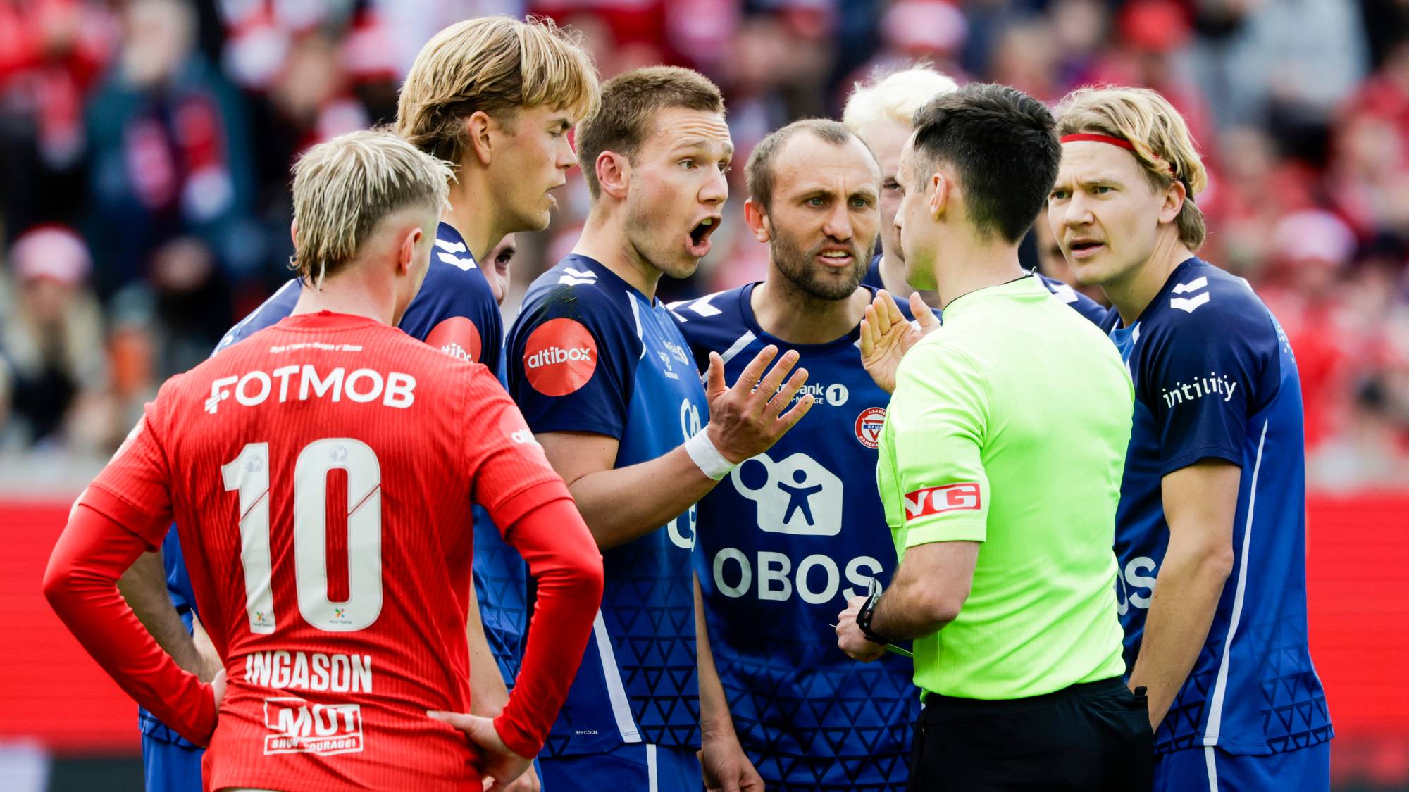 Fotballspillere er samlet rundt en dommer under en kamp. Spillere i blå og røde drakter er synlige. Publikum i bakgrunnen. Kampen spilles utendørs på en gressbane. (Bildebeskrivelsen er laget av en KI-tjeneste)