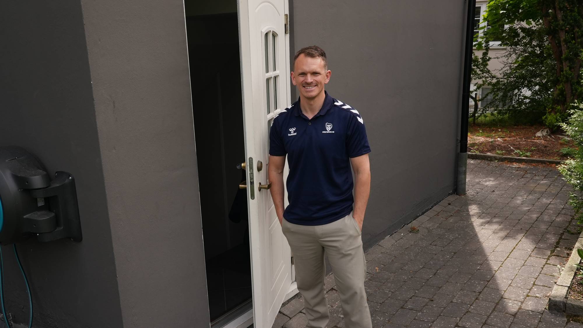 En mann står foran en mørk vegg og en åpen dør. Han er iført en mørkeblå pikétrøye med hvite detaljer og lyse bukser. Mannen smiler og har hendene i lommene. Utenfor er det en brosteinsbelagt gangvei og grøntområde. (Bildebeskrivelsen er laget av en KI-tjeneste)