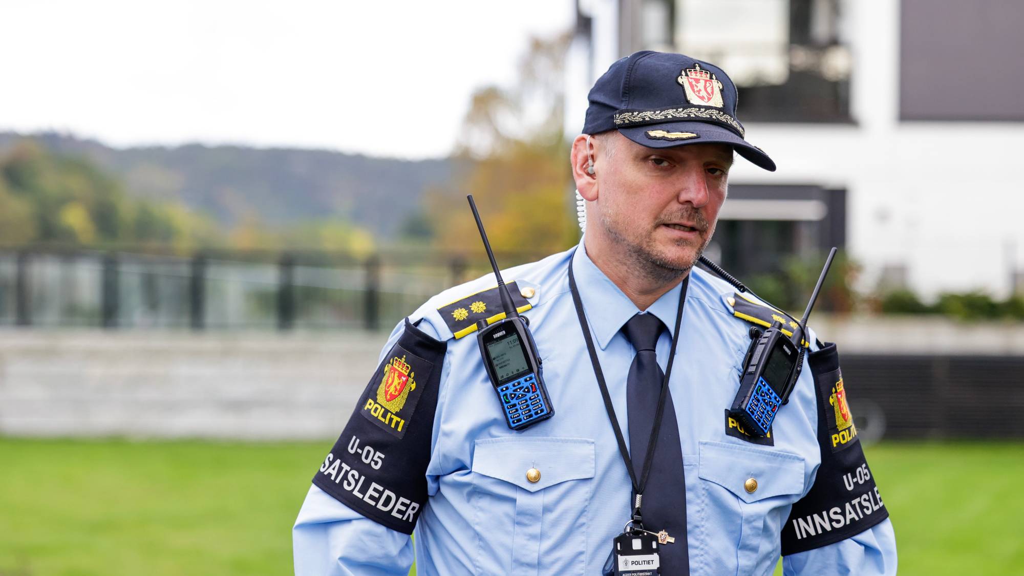Politimann med radiokommunikasjonsutstyr og ID-kort. Han er kledd i blå uniformskjorte med insignier og caps. I bakgrunnen sees en bygning og grøntområde. (Bildebeskrivelsen er laget av en KI-tjeneste)