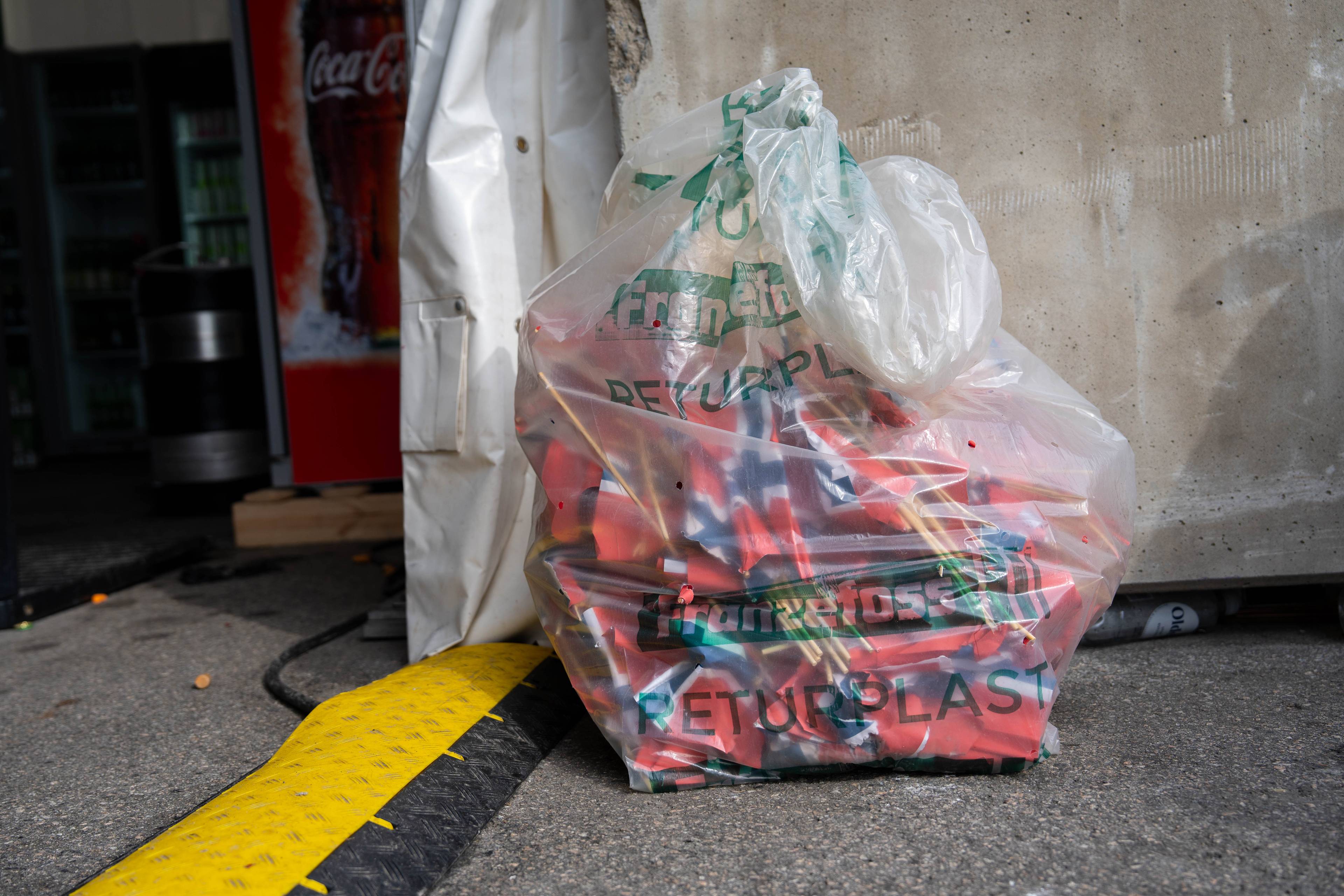 Ein gjennomsiktig plastsekk inneheld mange norske flagg. Sekken er merket med «RERETURNPLAST». Ved sidan av sekken står ein kvit presenning. I bakgrunnen er det ein Coca-Cola-kjøledisk. (Bildebeskrivelsen er laget av ein KI-teneste)