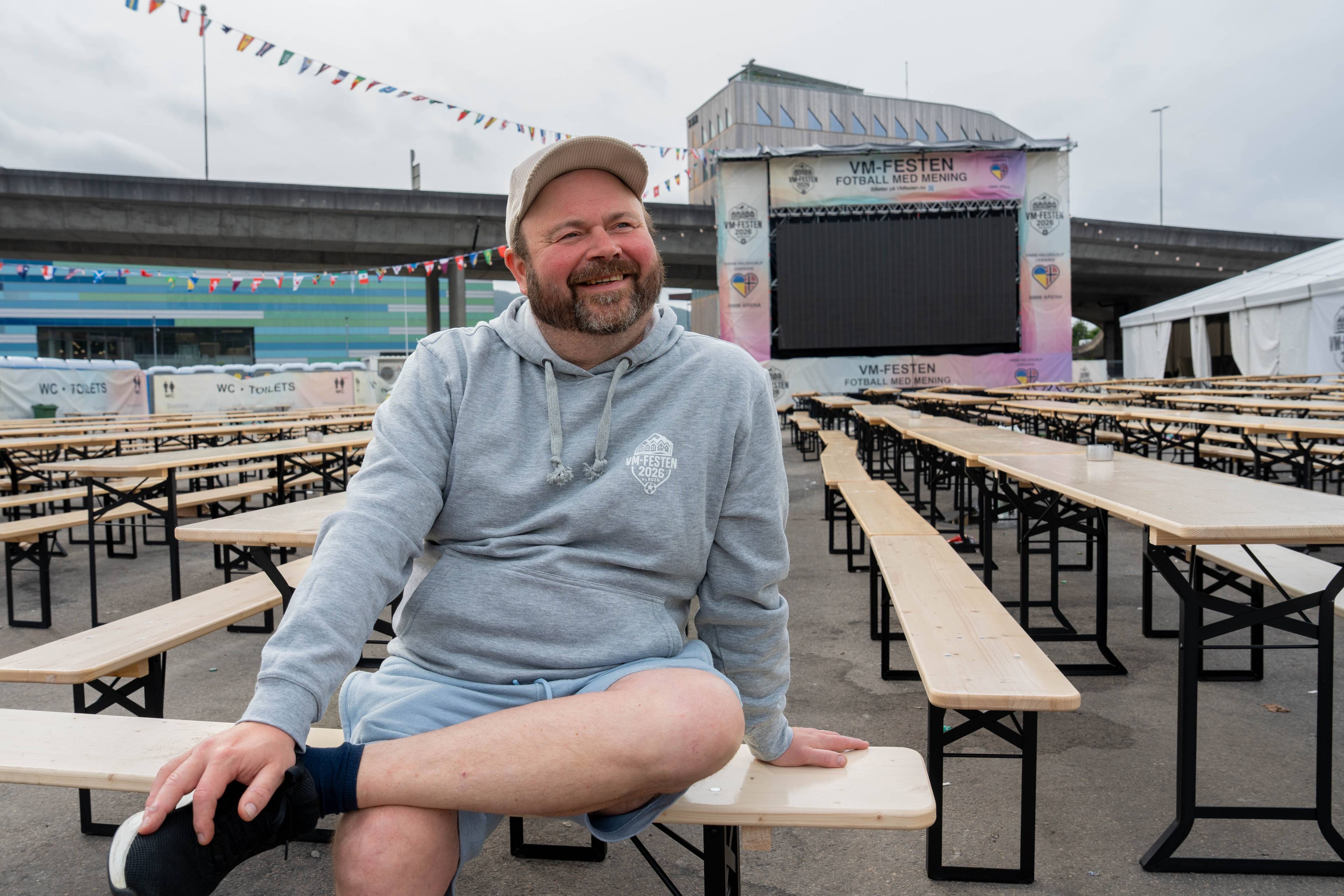 Ein mann sit på ein benk føre et stort antal tomme bord og benker. Bak ham er ein stor skjerm med teksten «VM-FESTEN FOTBALL MED MEINING». Området er pynta med flagg og det er fleire toalettbuer i bakgrunnen. (Bildebeskrivelsen er laget av ein KI-teneste)