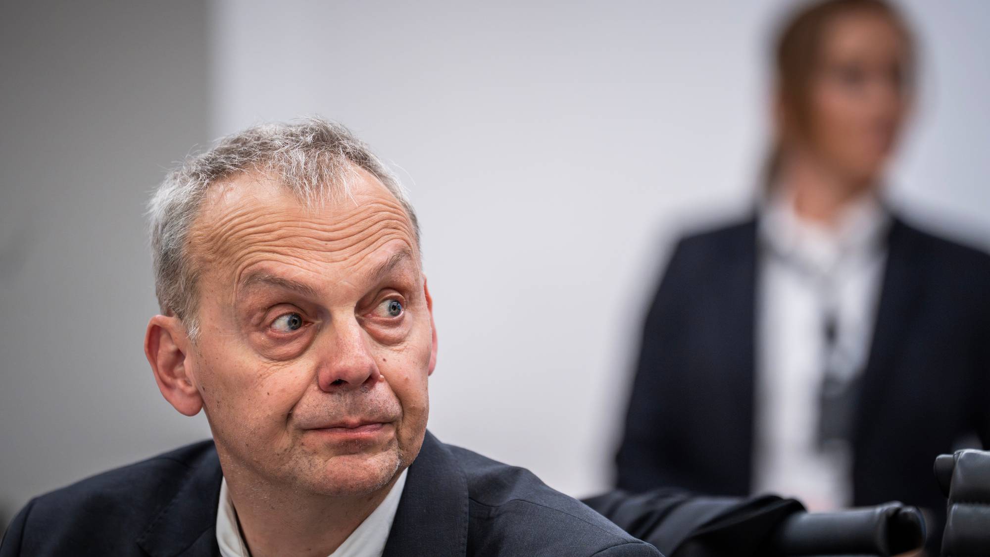 En mann i dress og slips sitter og ser til siden. I bakgrunnen sitter en annen person utydelig. (Bildebeskrivelsen er laget av en KI-tjeneste)