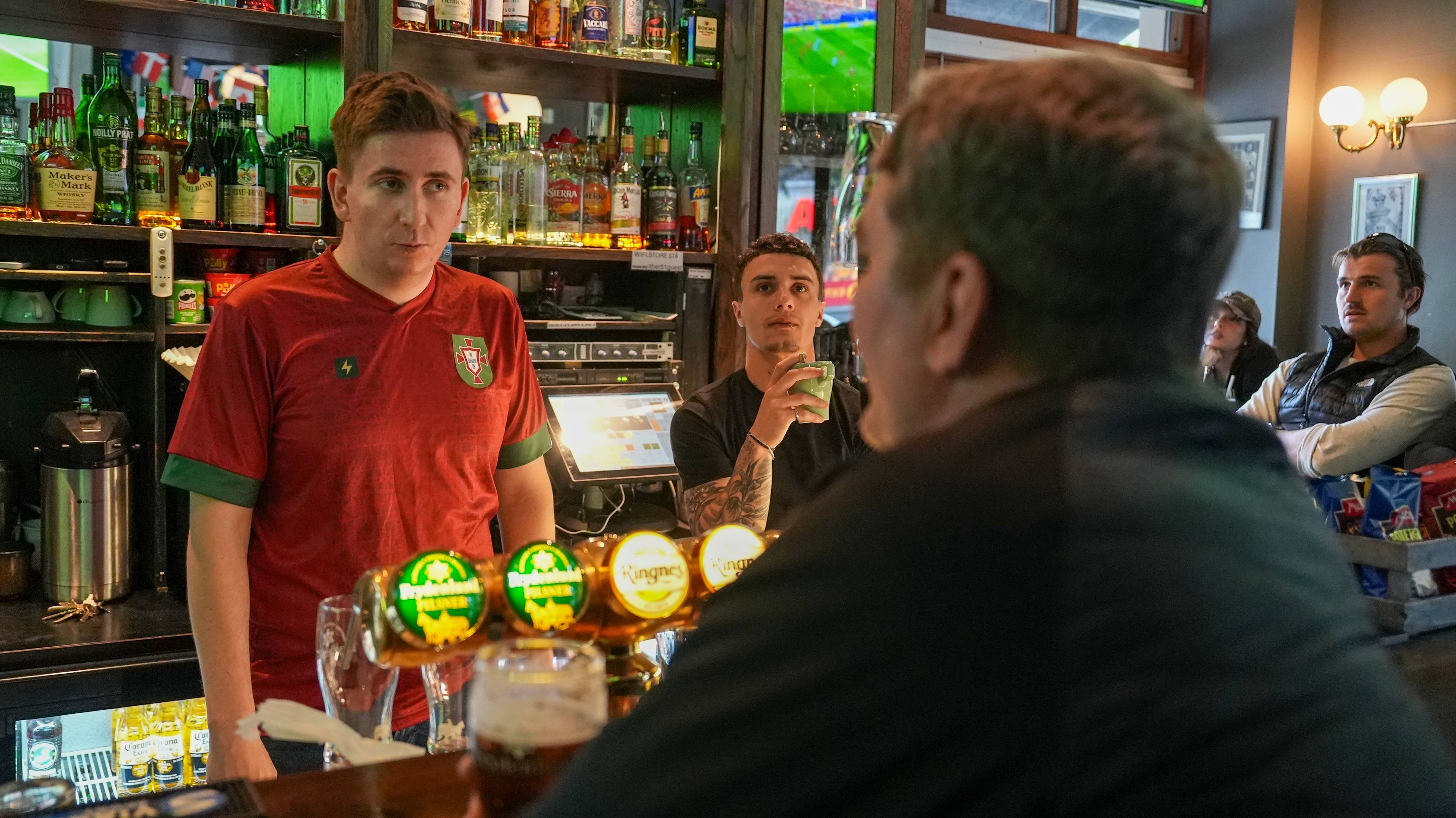En mann i en rød fotballtrøye står bak en bardisk. En annen mann drikker av en grønn kopp. Flere andre menn sitter ved bordet. Bildet er tatt inne i en pub. (Bildebeskrivelsen er laget av en KI-tjeneste)