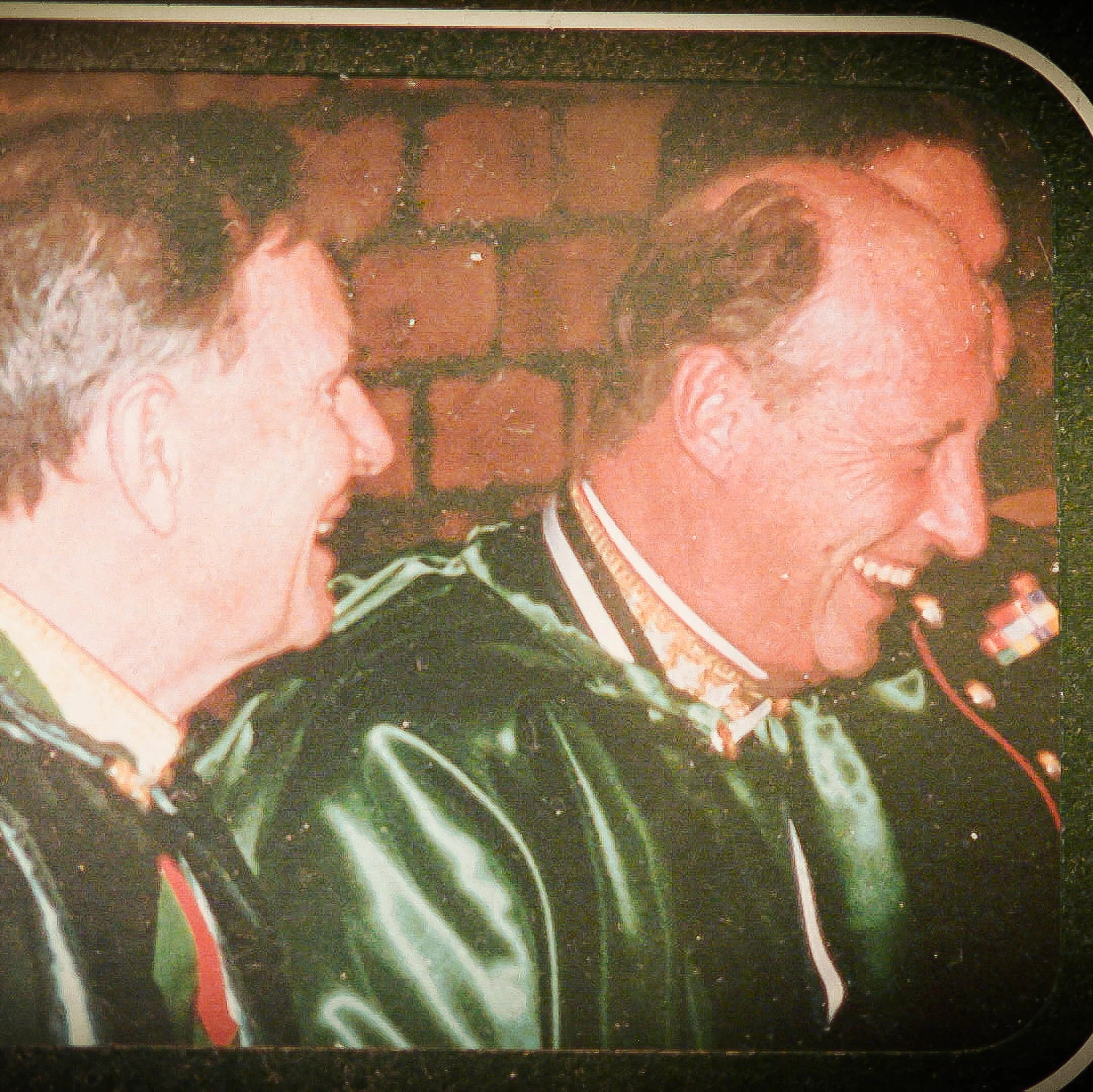 Kong Harald og en annen mann i mørke kapper ler sammen foran en teglsteinsvegg. (Bildebeskrivelsen er laget av en KI-tjeneste)