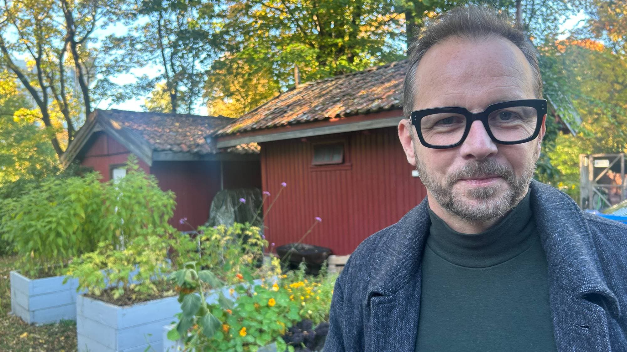 Portrett av en mann med briller foran en rød bod og plantekasser i en hage. (Bildebeskrivelsen er laget av en KI-tjeneste)