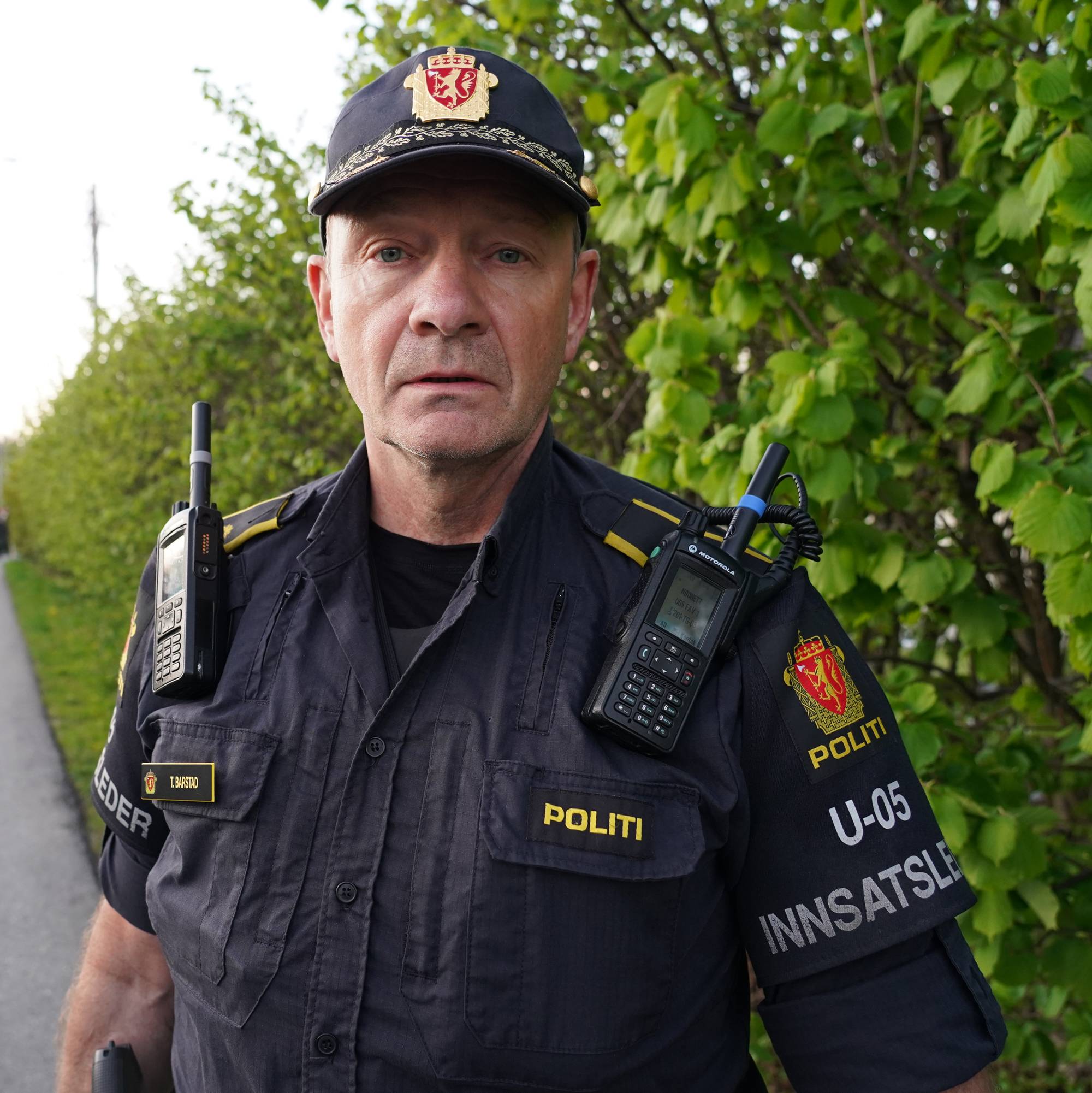 En politimann i uniform står vendt mot kameraet. Han har på seg en mørk uniformskjorte, caps og to radiokommunikatorer. I bakgrunnen er det en grønn hekk (Bildebeskrivelsen er laget av en KI-tjeneste)