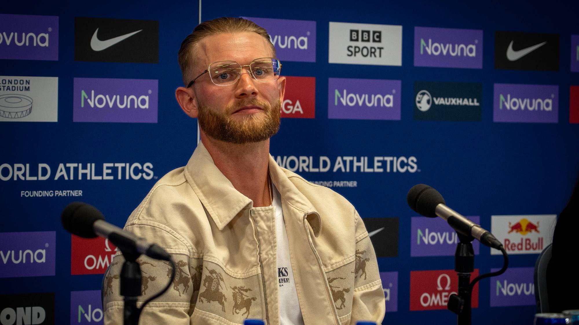 Josh Kerr sitter ved et bord med mikrofoner foran en World Athletics-vegg. (Bildebeskrivelsen er laget av en KI-tjeneste)
