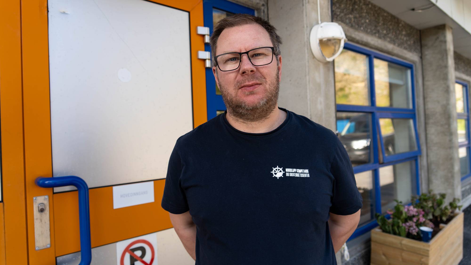 Mann med briller og t-skjorte står foran en bygningsinngang. (Bildebeskrivelsen er laget av en KI-tjeneste)