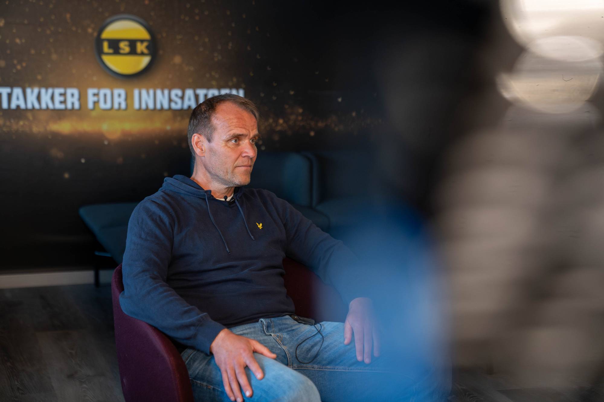 En mann sitter på en mørk sofa foran en vegg med ovale portrettbilder. Bildene viser ansiktene til flere personer i rammer. Bakgrunnen er en lyseblå vegg.