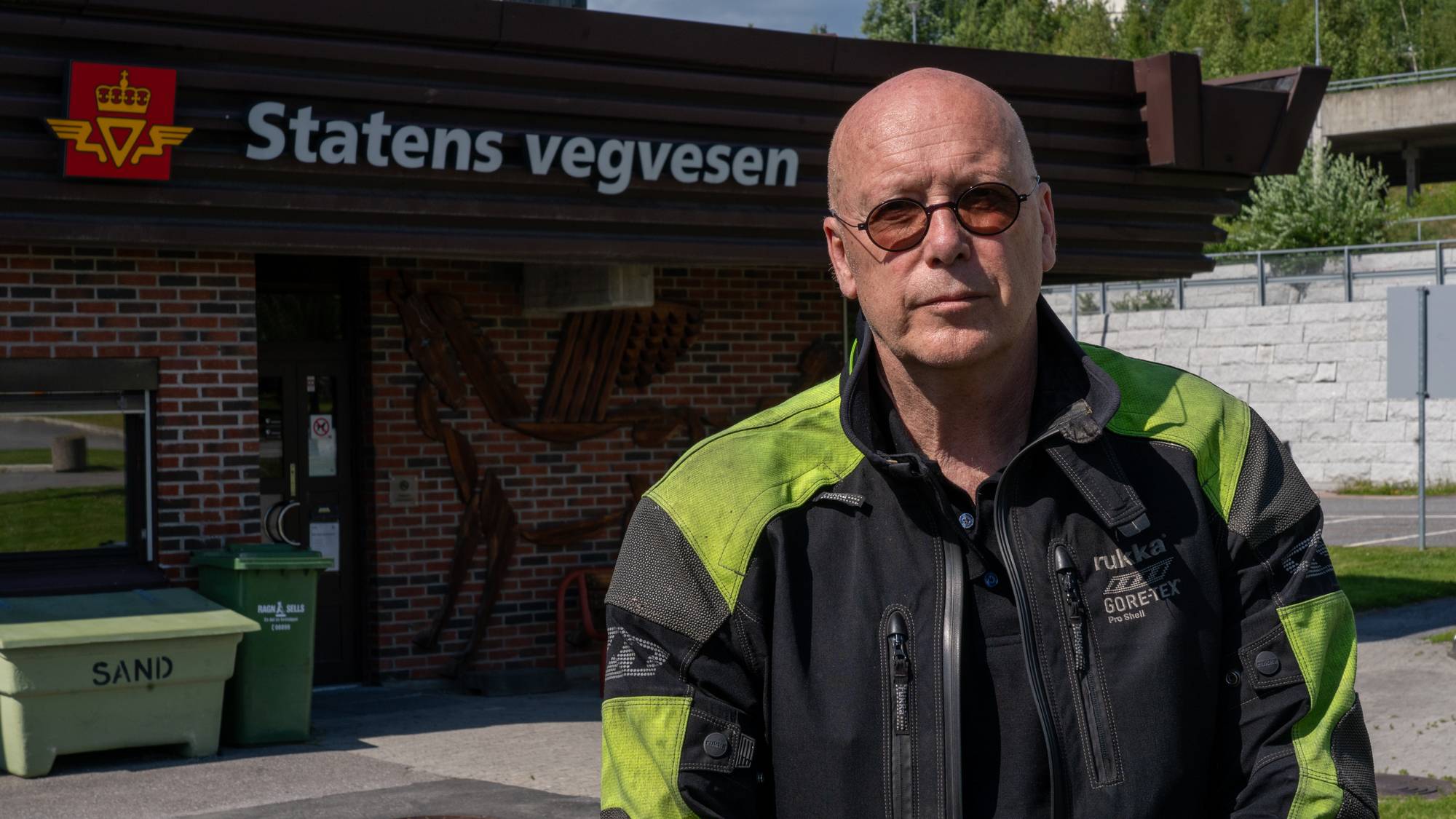 En mann med solbriller og motorsykkeljakke står utenfor en bygning med logo og teksten "Statens vegvesen" på.