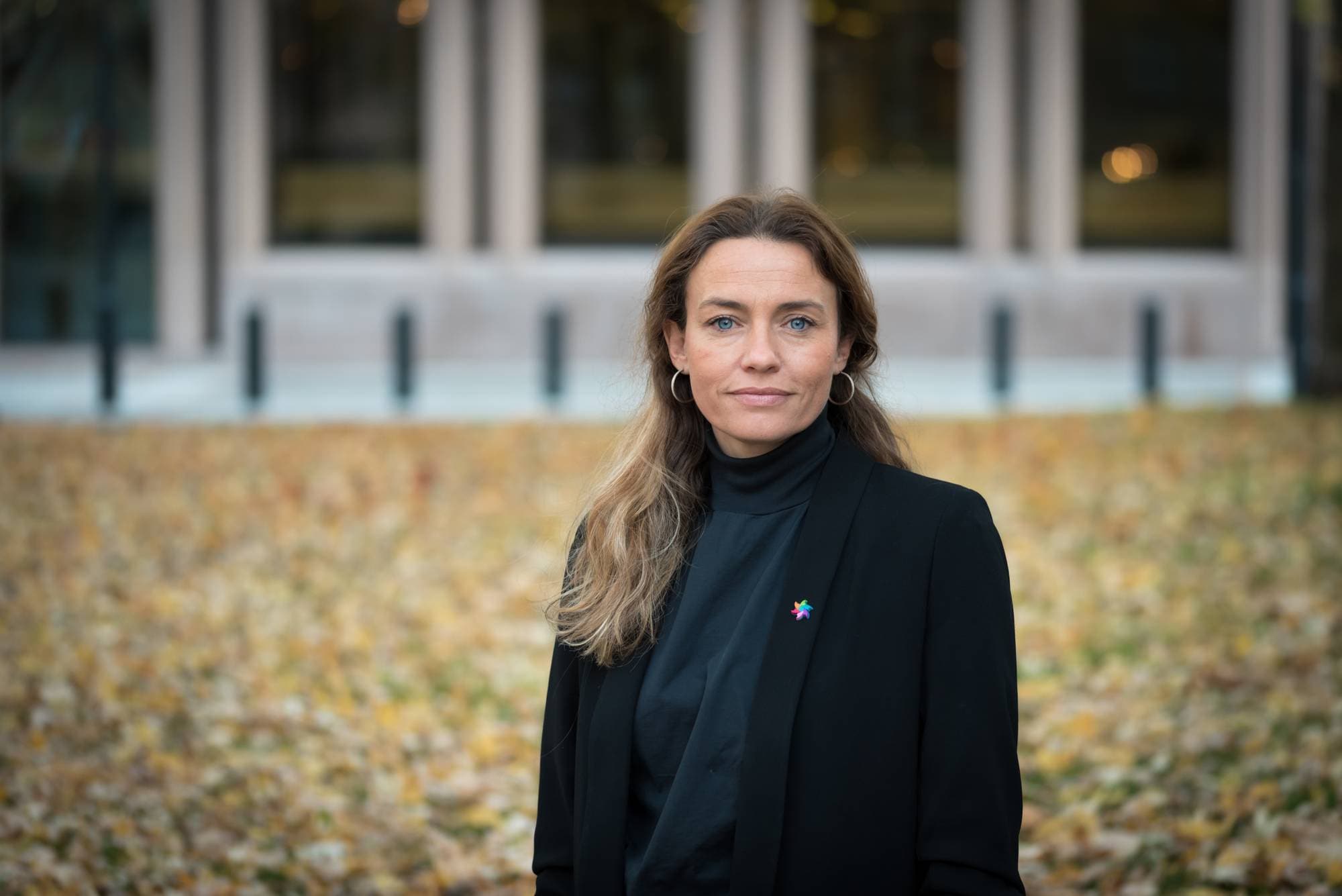 En kvinne står i en offentlig park med høstløv på bakken. Hun har langt, bølgete hår og iført en svart dressjakke over en mørk genser. I bakgrunnen kan man skimte bygninger med store vinduer.