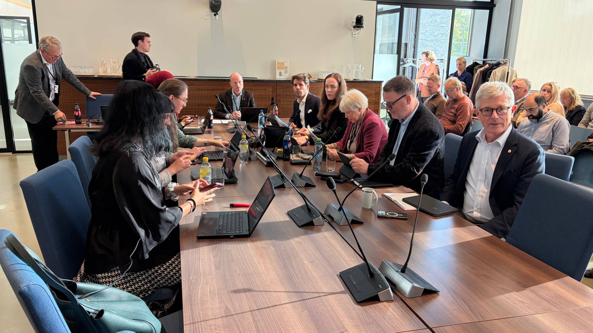 En gruppe mennesker sitter rundt et langt møtebord i et lyst rom. De bruker laptoper og mikrofoner. (Bildebeskrivelsen er laget av en KI-tjeneste)