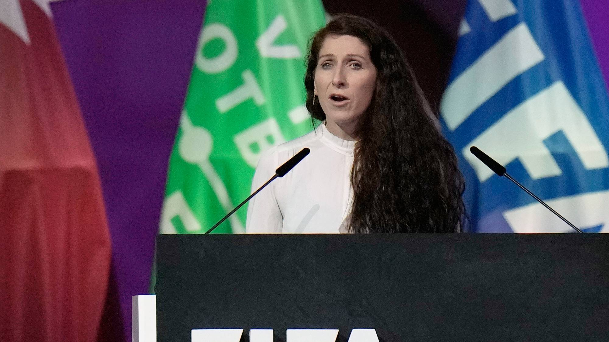 Kvinne med langt mørkt hår taler fra en talerstol med FIFA-logo foran flagg. (Bildebeskrivelsen er laget av en KI-tjeneste)