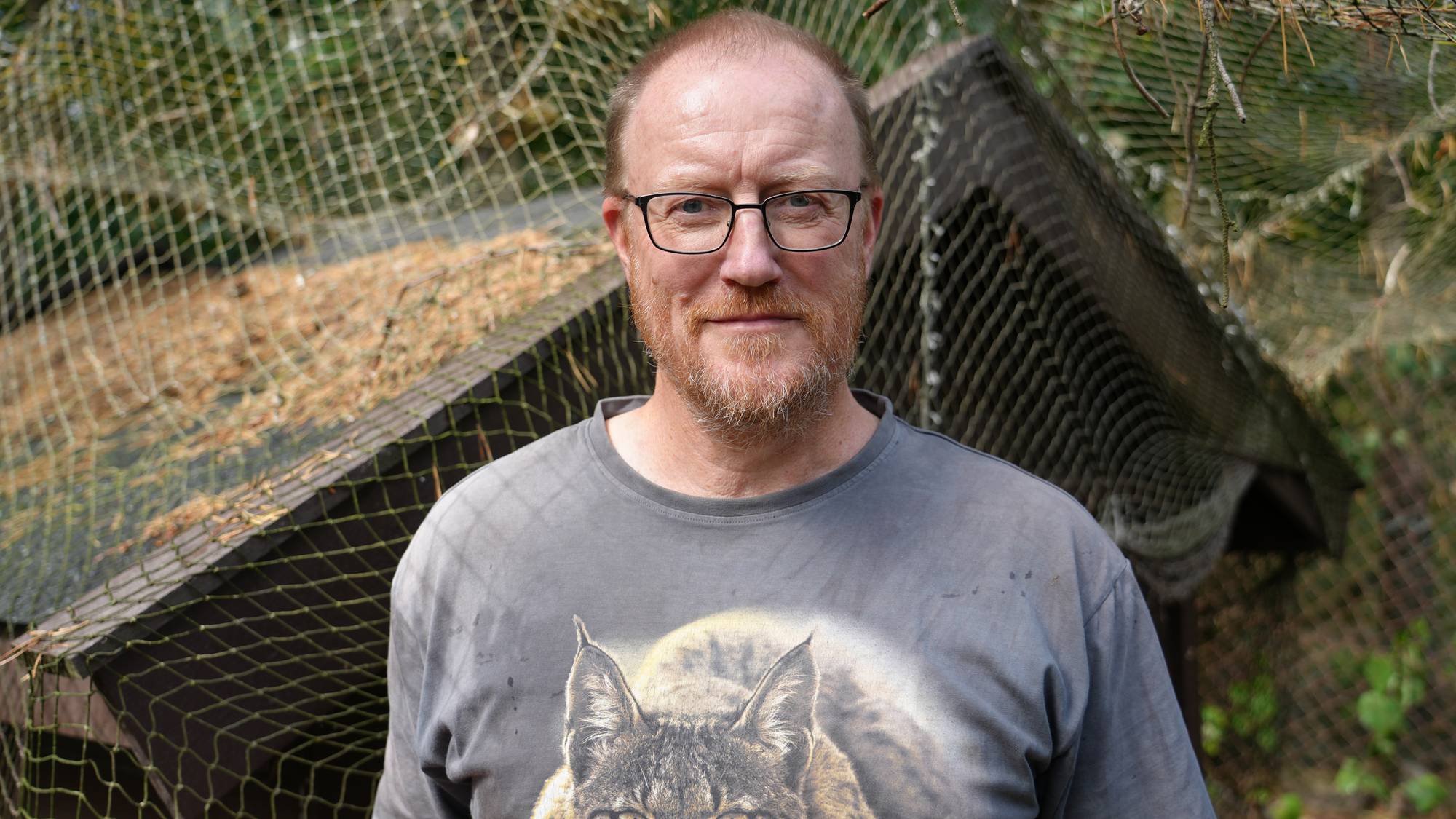 En mann som står foran et hønsehus med netting. I bakgrunnen er det skog.