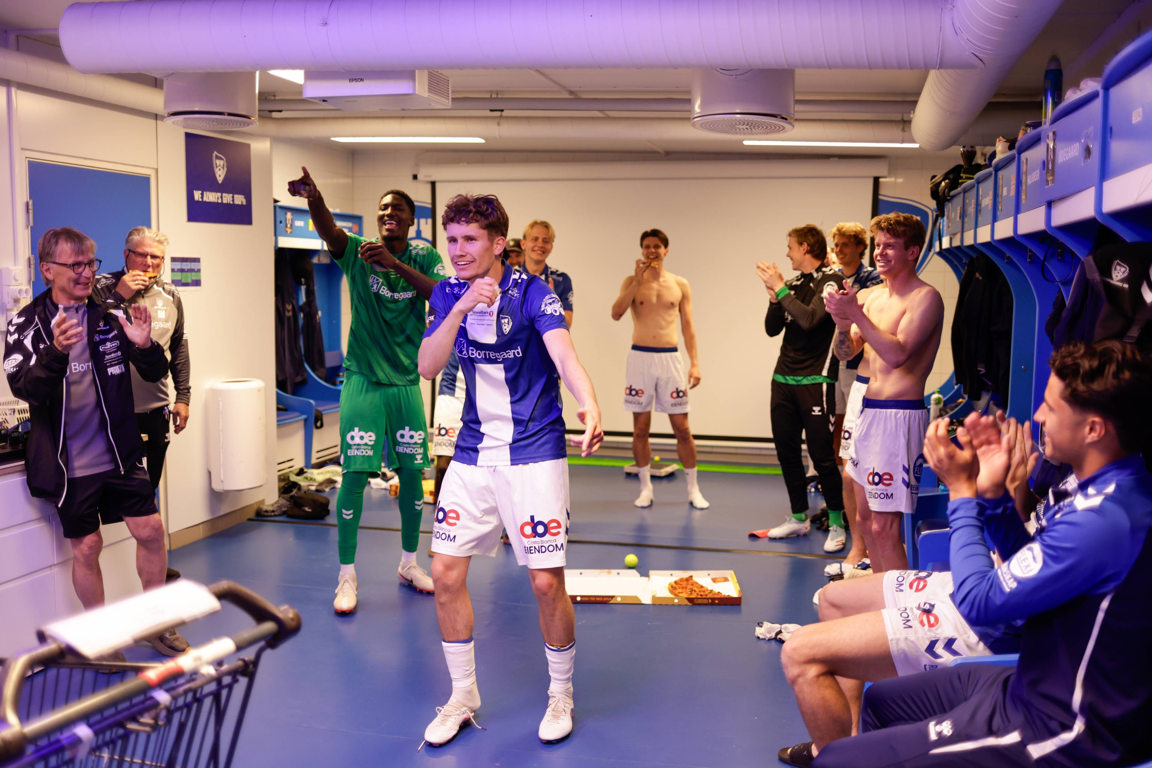 En gruppe fotballspillere feirer i garderoben. n spiller i blåhvit drakt klapper mot kamera.Andre spillere i grønne og blå drakter er også til stede. En person i jakke klapper.I bakgrunnen er det blå skap og utstyr. En pizza ligger på gulvet. (Bildebeskrivelsen er laget av en KI-tjeneste)
