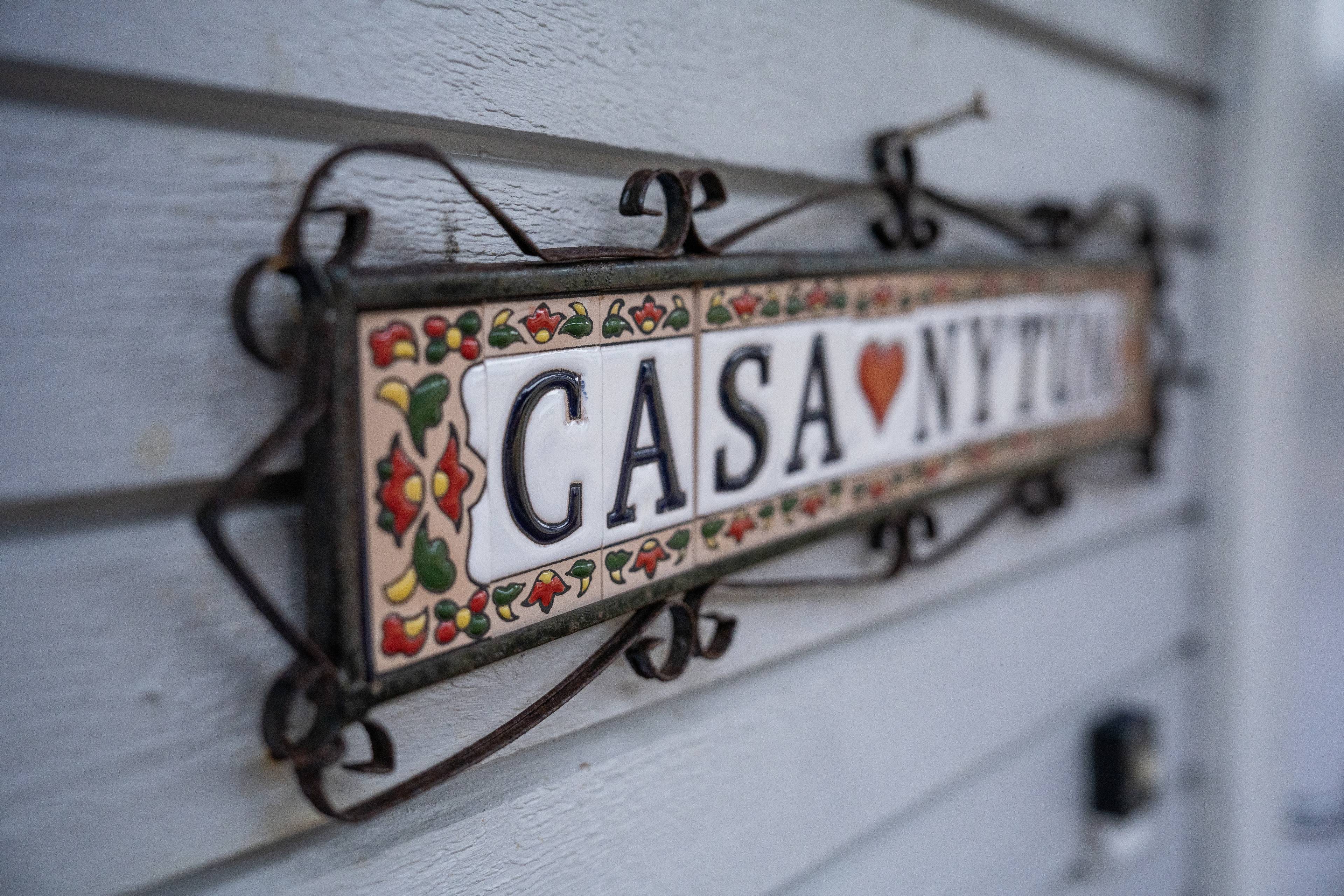 Et hus-skilt med teksten "CASA NYTUN" påmontert en hvit trevegg. Skiltet er dekorert med blomstermotiv og en hjerteformet rød figur mellom ordene "CASA" og "NYTT". Skiltet henger på veggen med et utsmykket jernfeste. En liten, mørk dørklokke er synlig på veggen til høyre for skiltet. (Bildebeskrivelsen er laget av en KI-tjeneste)