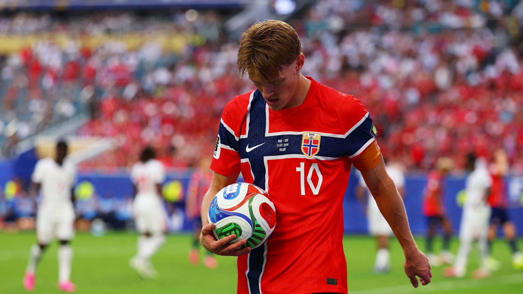 Martin Ødegaard i norsk landslagsdrakt holder en fotball på en bane med publikum i bakgrunnen. (Bildebeskrivelsen er laget av en KI-tjeneste)