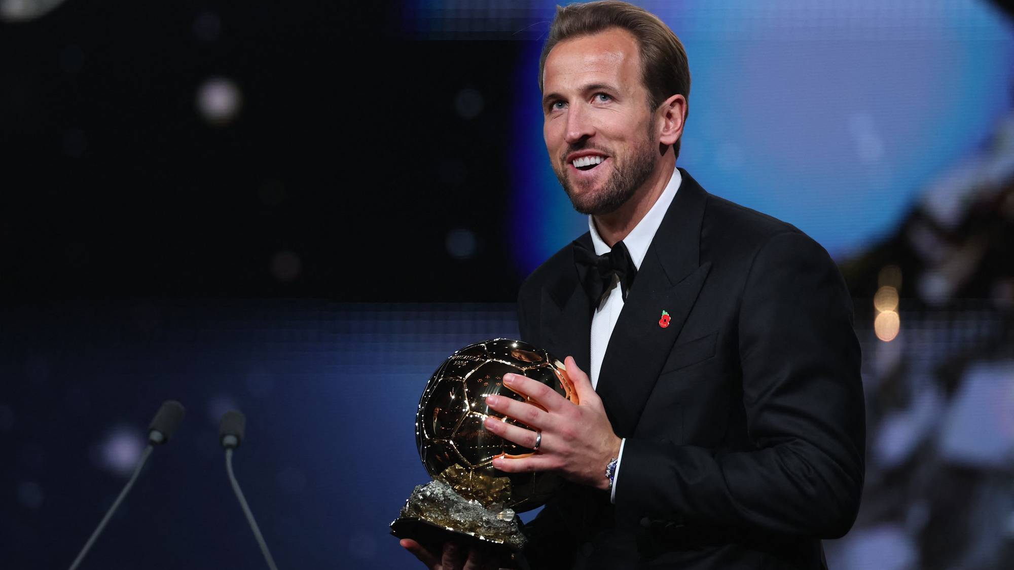 Harry Kane smiler mens han holder en fotballtrofé på en scene foran en mørk bakgrunn. (Bildebeskrivelsen er laget av en KI-tjeneste)