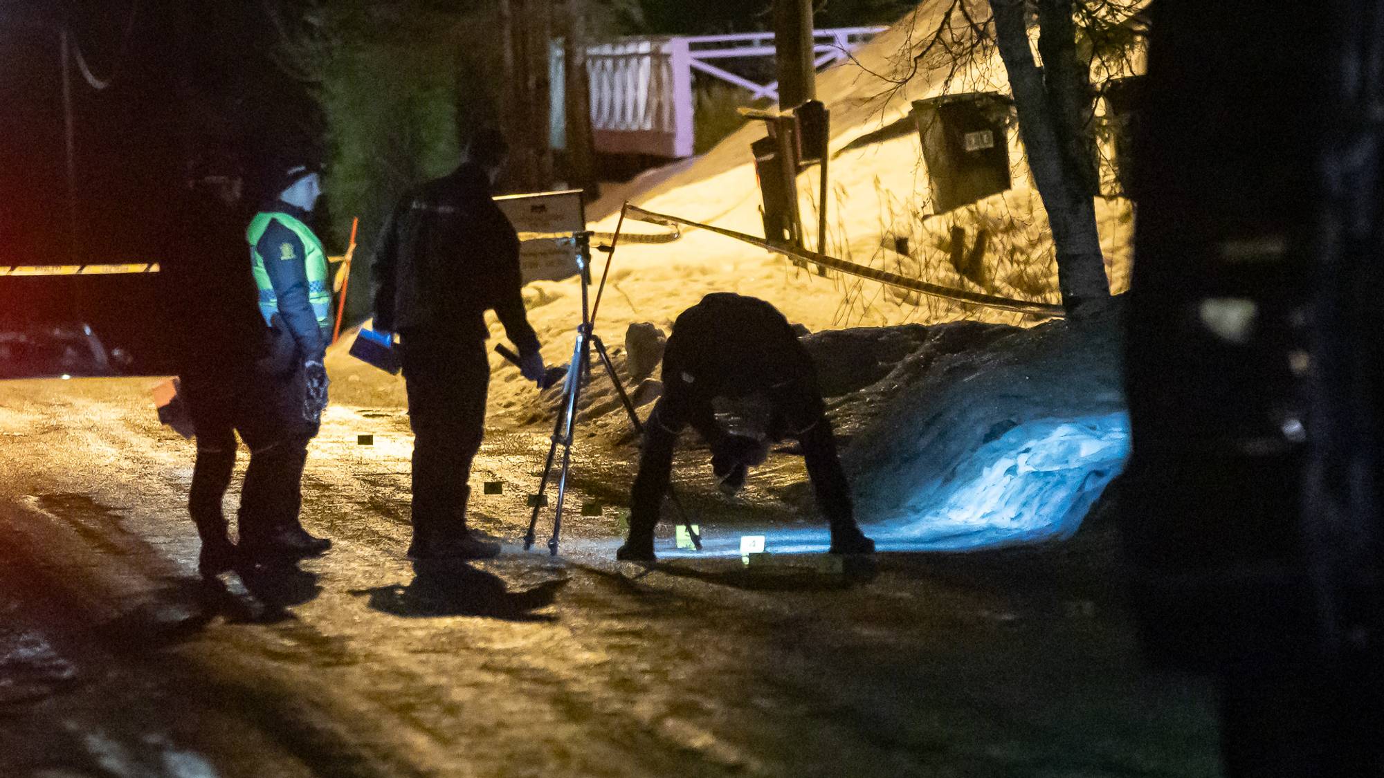 Politiet etterforsker en åstedsscene i en snødekt gate om natten. To personer i politiuniform undersøker bakken, mens en tredje bøyer seg for å samle bevis. Området er delvis opplyst, med snø og en traktorvei synlig i bakgrunnen. Deler av scenen er avsperret med politibånd. (Bildebeskrivelsen er laget av en KI-tjeneste)