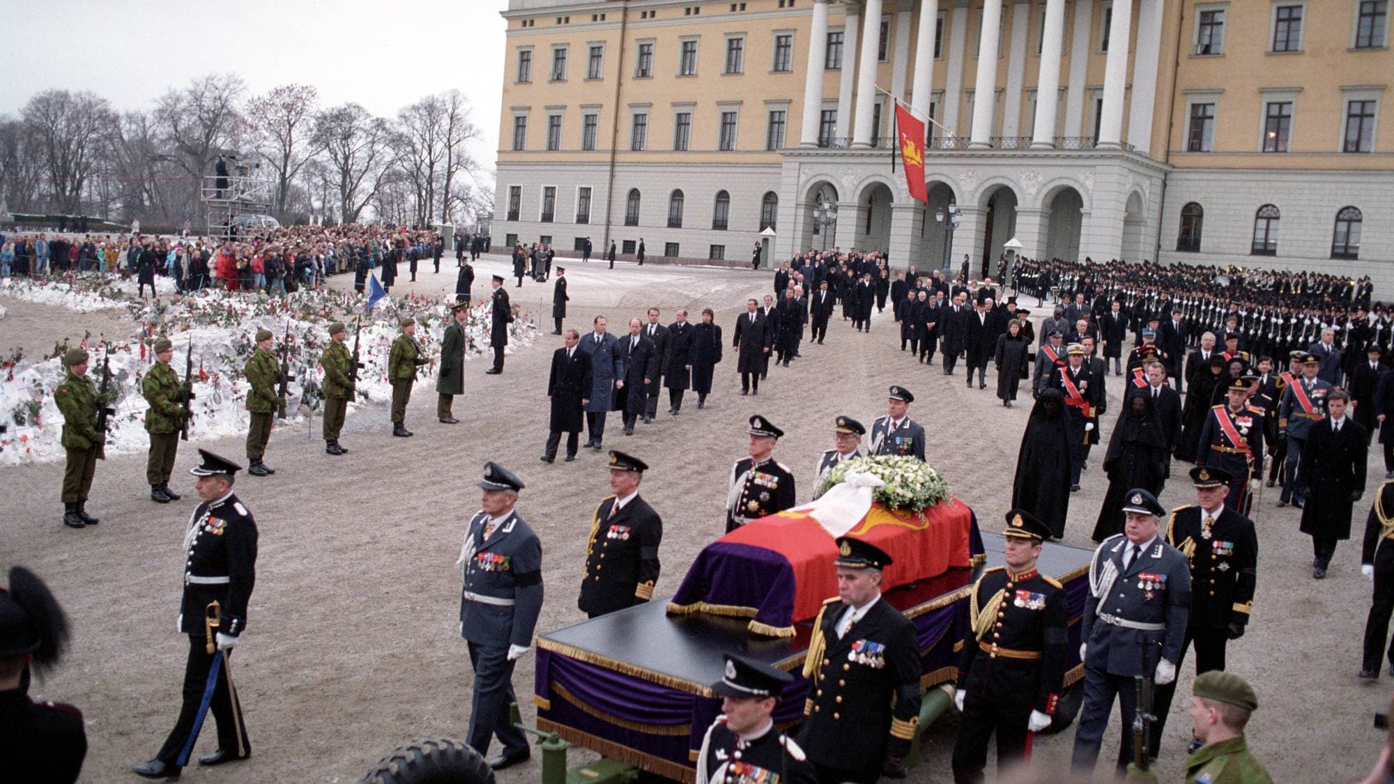 Kong Olavs båre ligger på en kanonlavett som blir trukket av et militærkjøretøy, og bak følger prosesjonen som går fra Slottet til begravelsesseremonien i Oslo domkirke 30. januar 1991
