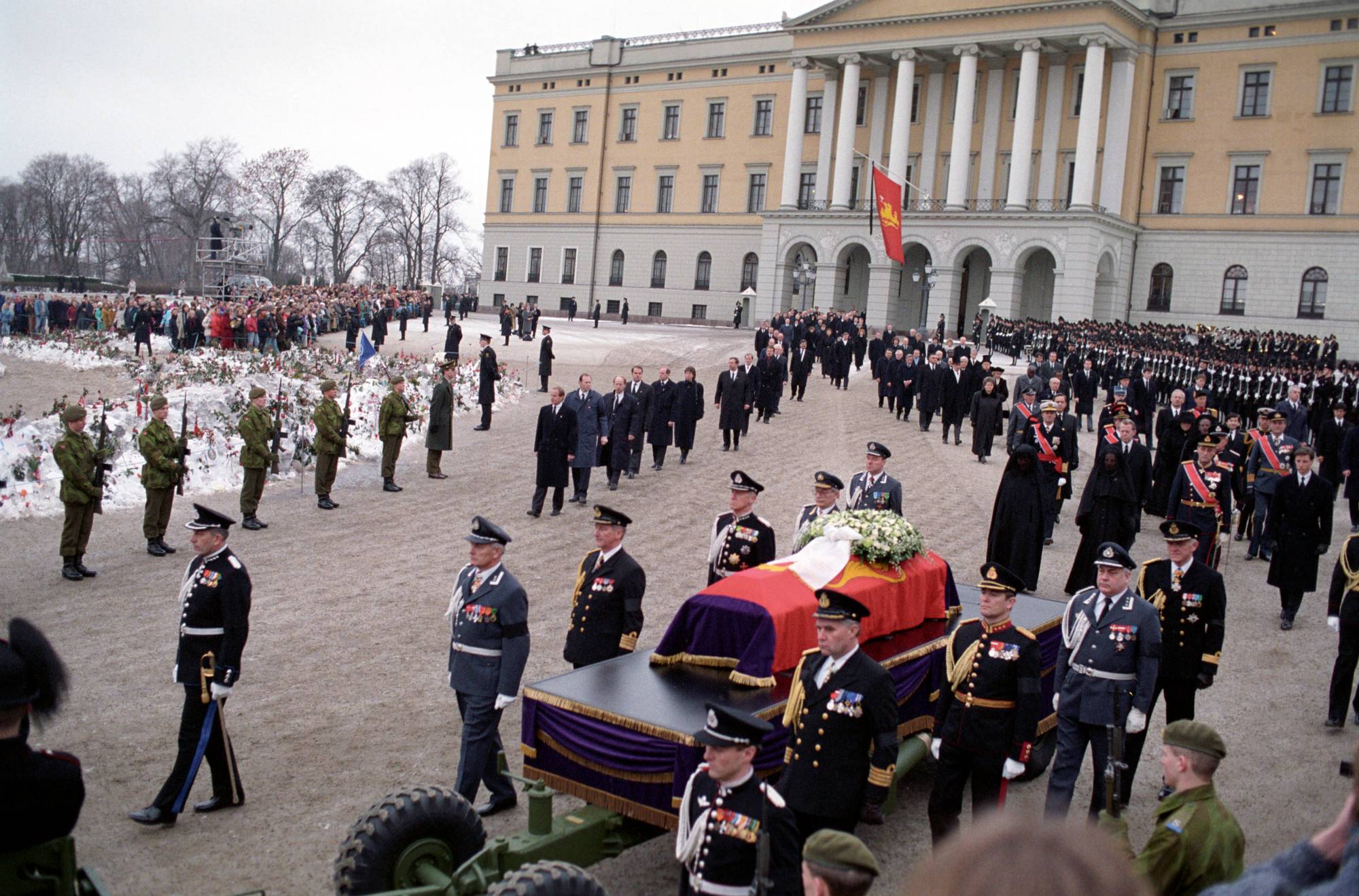 Kong Olavs båre ligger på en kanonlavett som blir trukket av et militærkjøretøy, og bak følger prosesjonen som går fra Slottet til begravelsesseremonien i Oslo domkirke 30. januar 1991