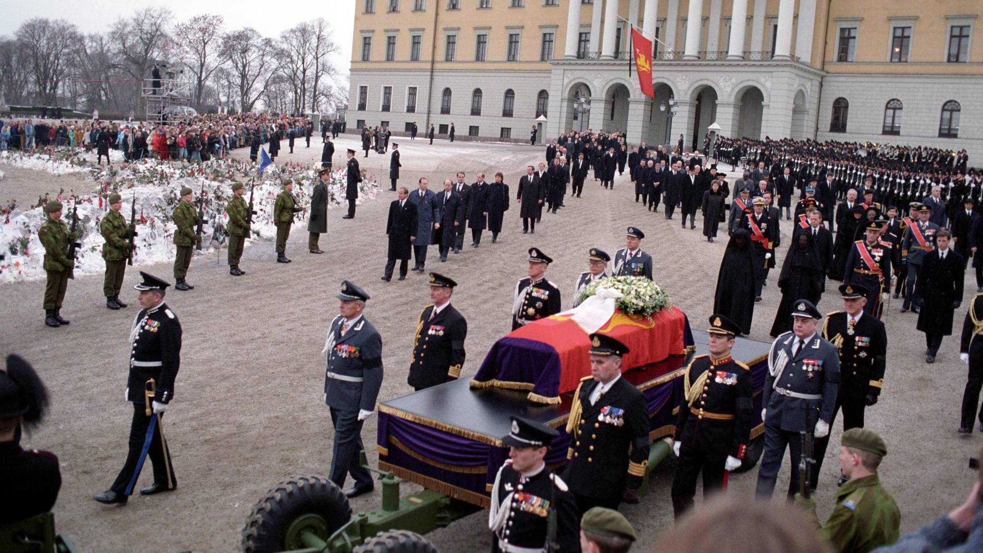 Kong Olavs båre ligger på en kanonlavett som blir trukket av et militærkjøretøy, og bak følger prosesjonen som går fra Slottet til begravelsesseremonien i Oslo domkirke 30. januar 1991