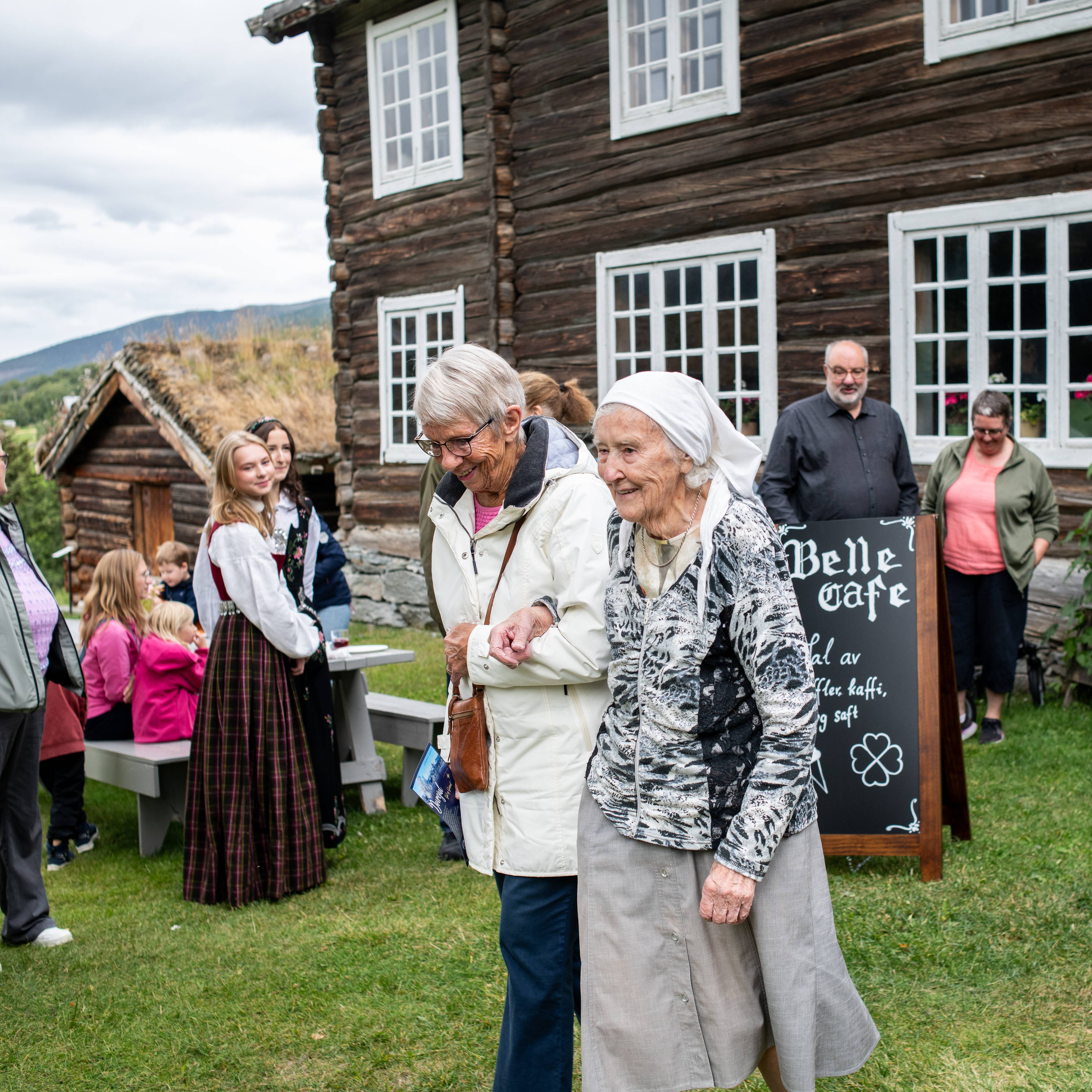 To eldre kvinner går sammen foran et eldre tømmerhus. Flere mennesker er samlet i bakgrunnen.