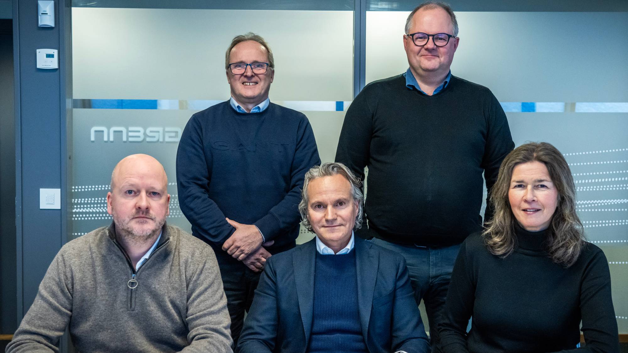 Fem personer sitter og står sammen foran en glassvegg på et kontor. (Bildebeskrivelsen er laget av en KI-tjeneste)