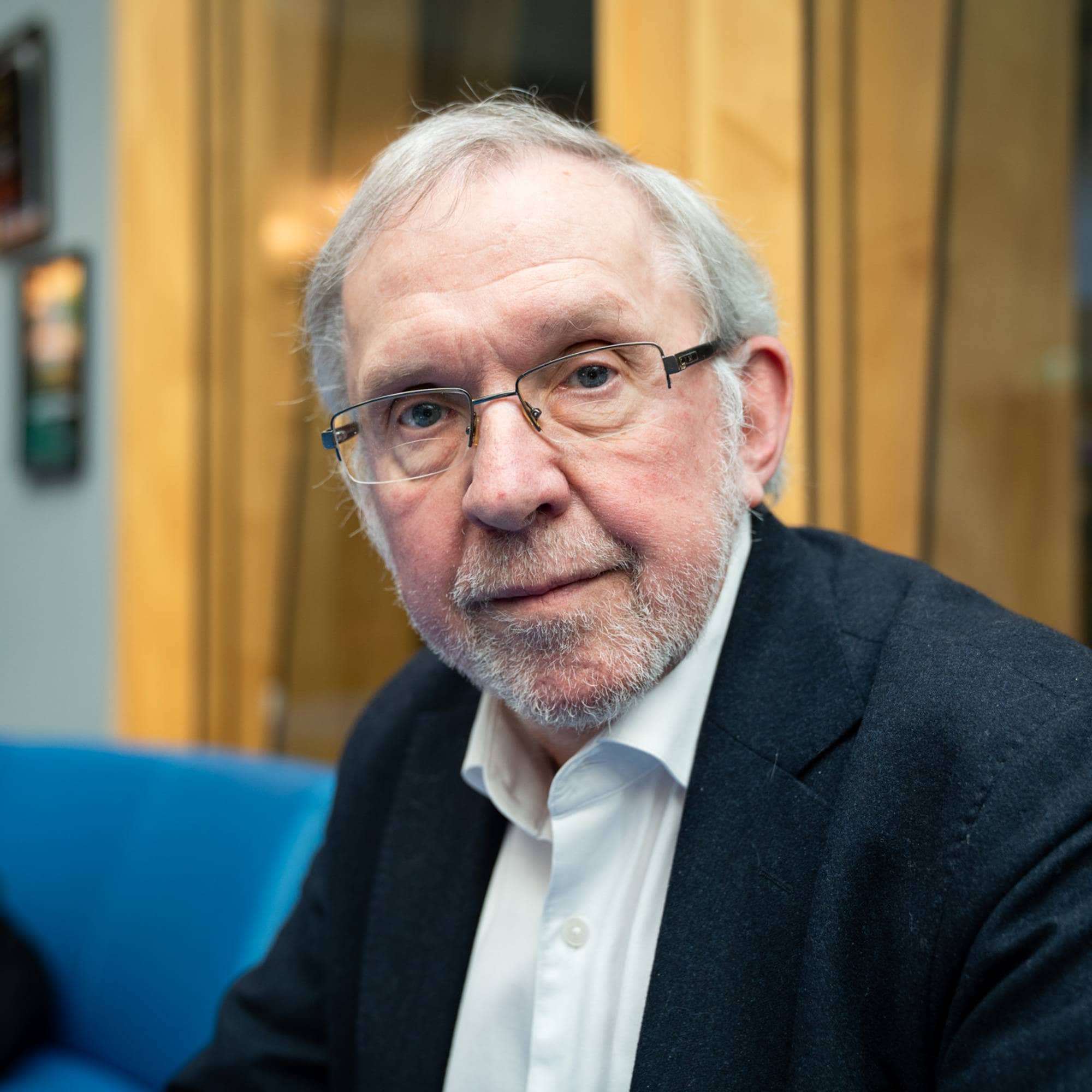 Mann med grått hår og skjegg og briller i dress på blå sofa