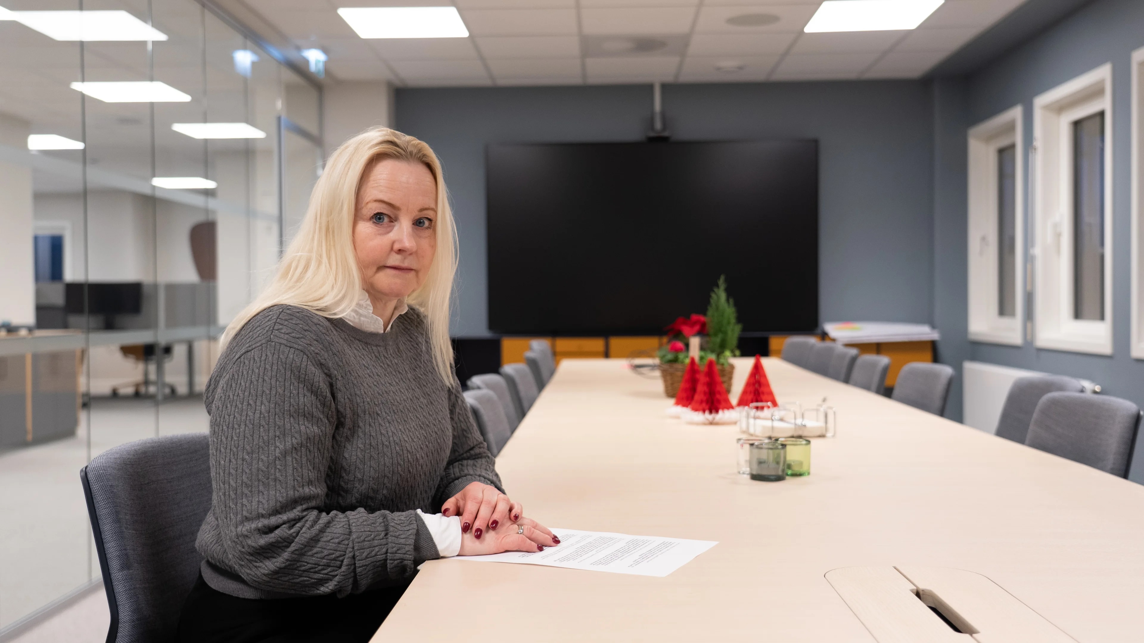 En kvinne sitter ved et langt konferansebord i et moderne møterom. Hun har på seg en grå genser og ser ned på et papir som ligger foran henne. På bordet er det dekorasjoner, inkludert små, røde juletreprydnader og lys. I bakgrunnen er det en stor skjerm og glassvegger som gir innsyn til resten av rommet. (Bildebeskrivelsen er laget av en KI-tjeneste)