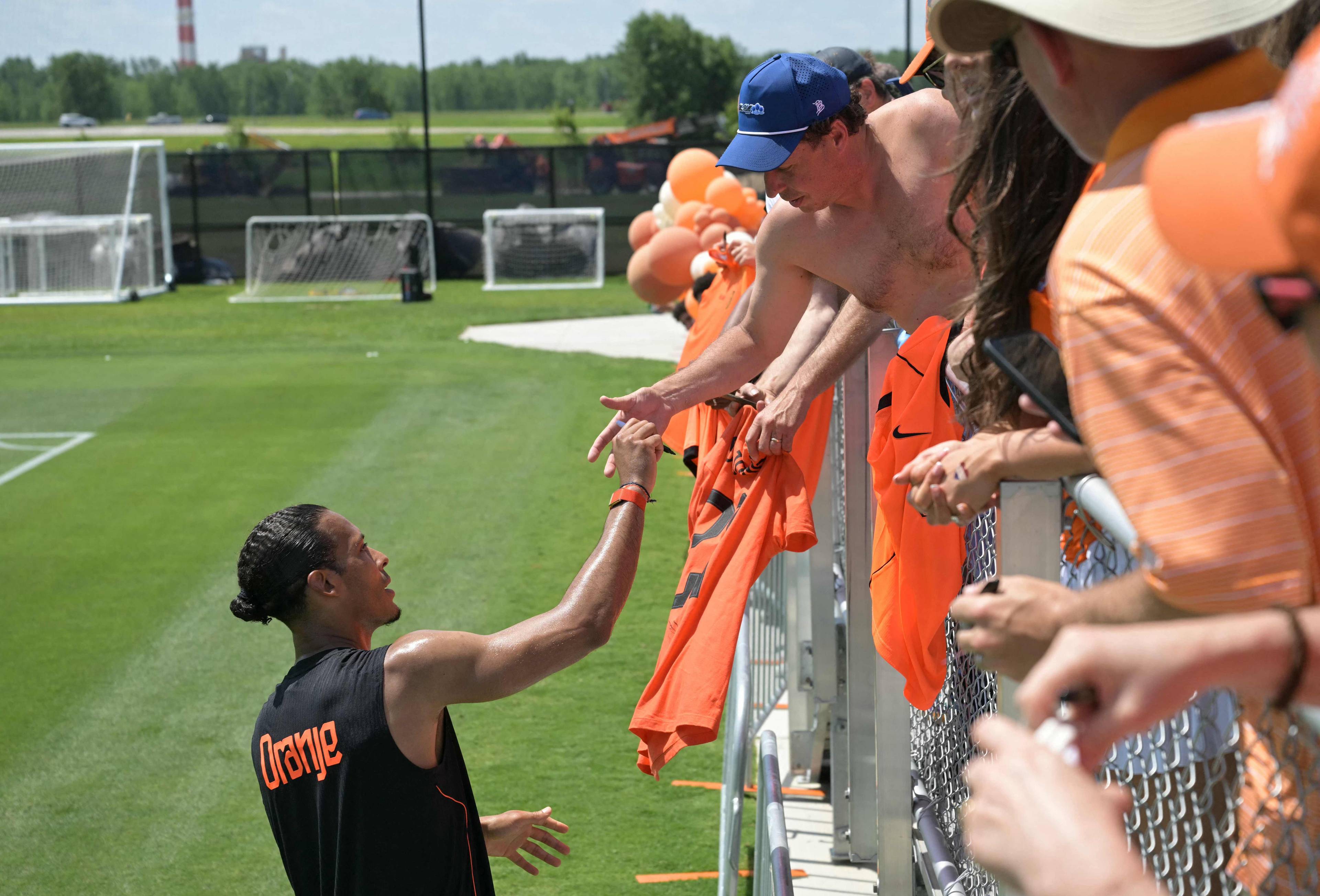 En fotballspiller i svart drakt med teksten "Orange" på gir en t-skjorte til supportere ved et gjerde. Supporterne, flere kledd i oransje, ser ut til å ta imot t-skjorten. Bildet er tatt på en gressbane med fotballmål i bakgrunnen. (Bildebeskrivelsen er laget av en KI-tjeneste)