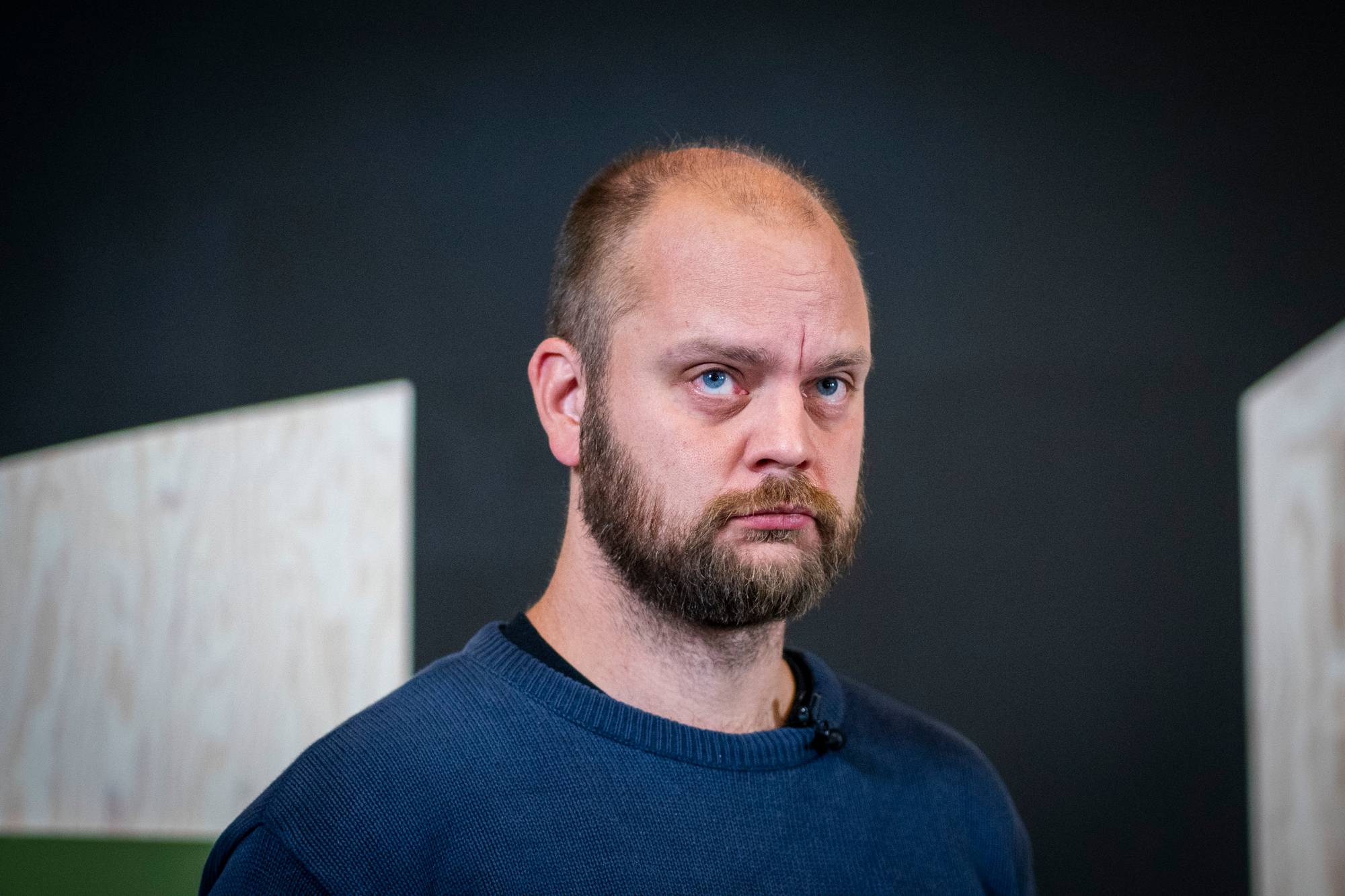 Mann med kort hår og skjegg ser mot kamera. Han har på seg en mørkeblå genser. Bakgrunnen er mørk med et lyst panel til venstre. (Bildebeskrivelsen er laget av en KI-tjeneste)