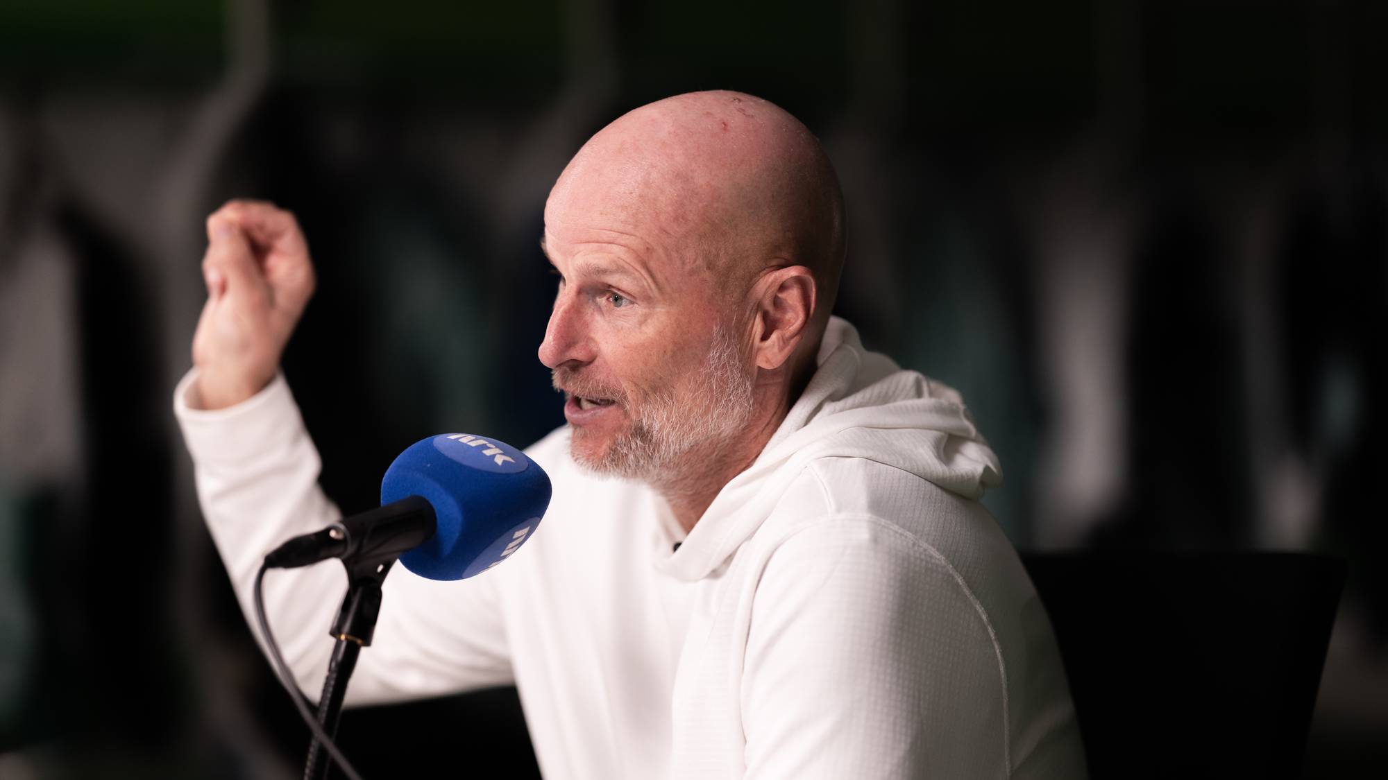 Ståle Solbakken snakker i en NRK-mikrofon med løftet hånd. (Bildebeskrivelsen er laget av en KI-tjeneste)