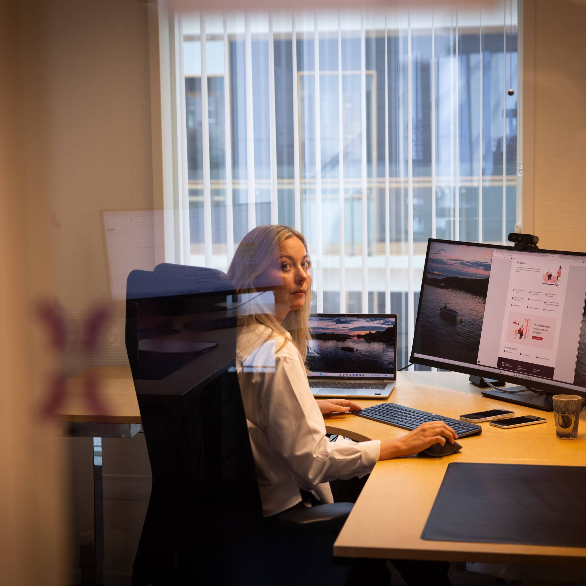 En kvinne sitter ved en pult hvor hun jobber på en pc. Hun ser i kamera over skulderen sin