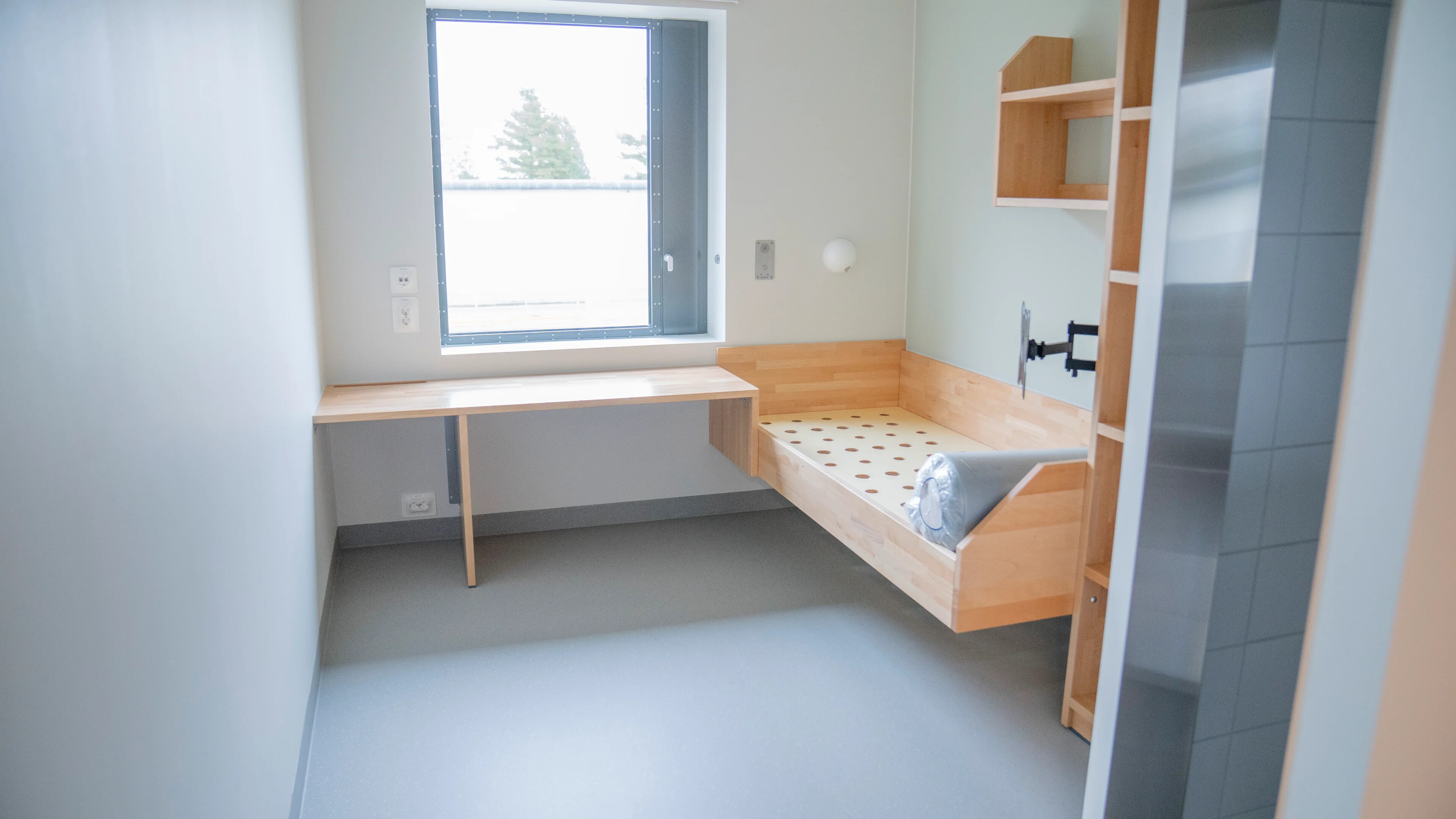 En moderne fengselscelle med seng, skrivebord og hylle. Lyst rom med vindu.