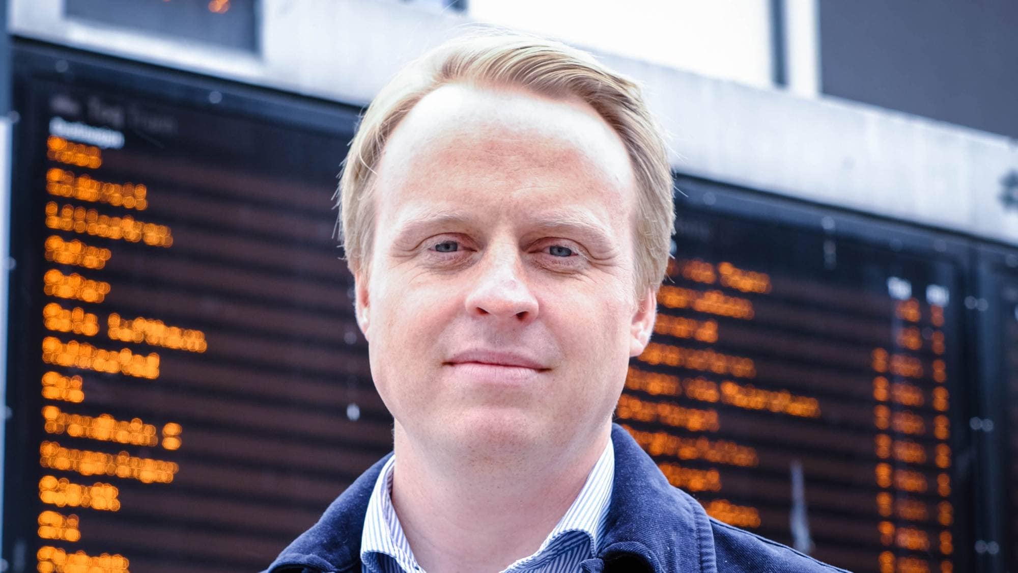 En mann med kort, lyst hår ser inn i kamera. Han har på seg en lys skjorte og blå jakke. 
