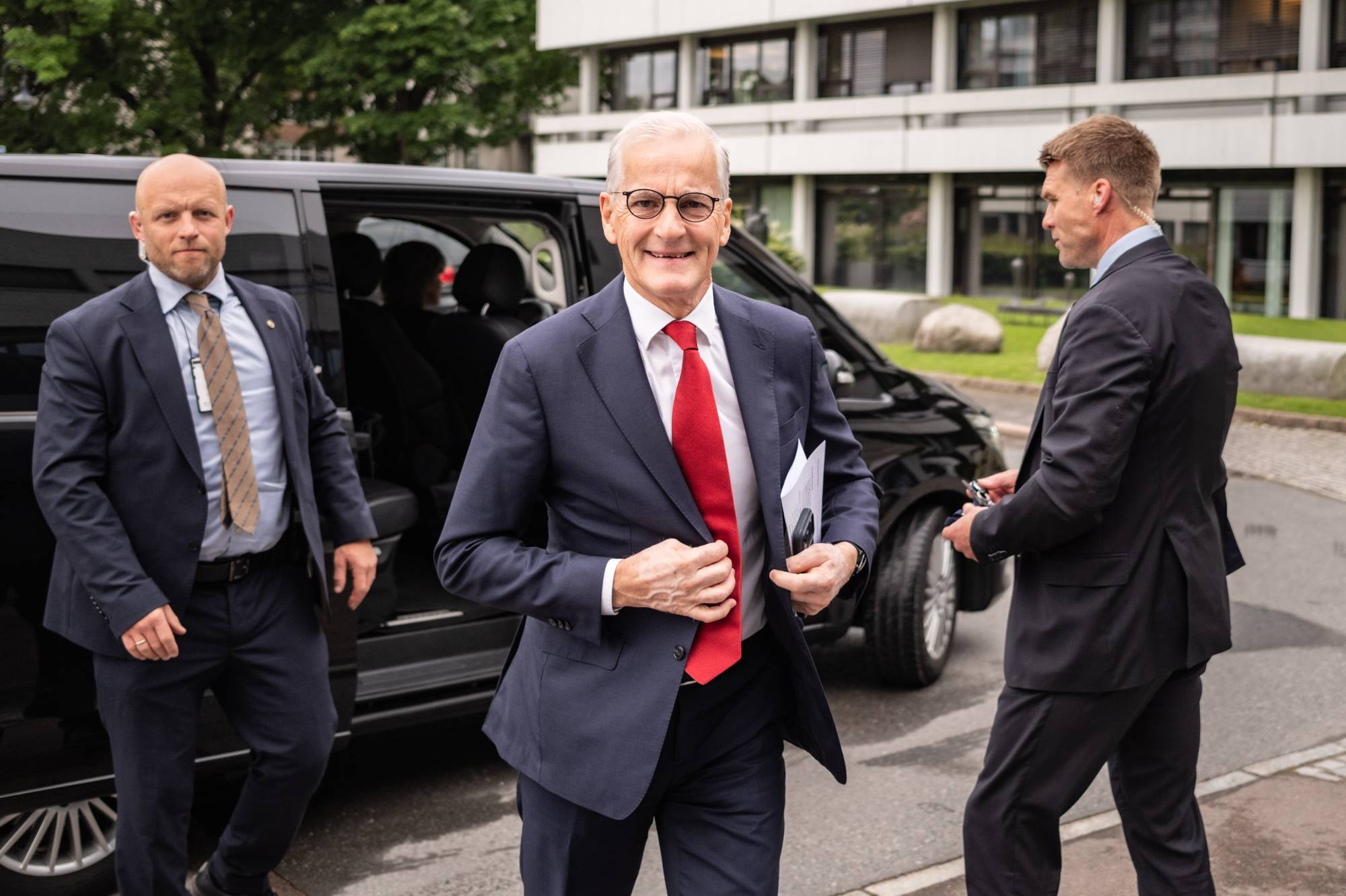Mann i dress og slips kommer ut av sort bil. En mann med dress på hver av sidene hans.