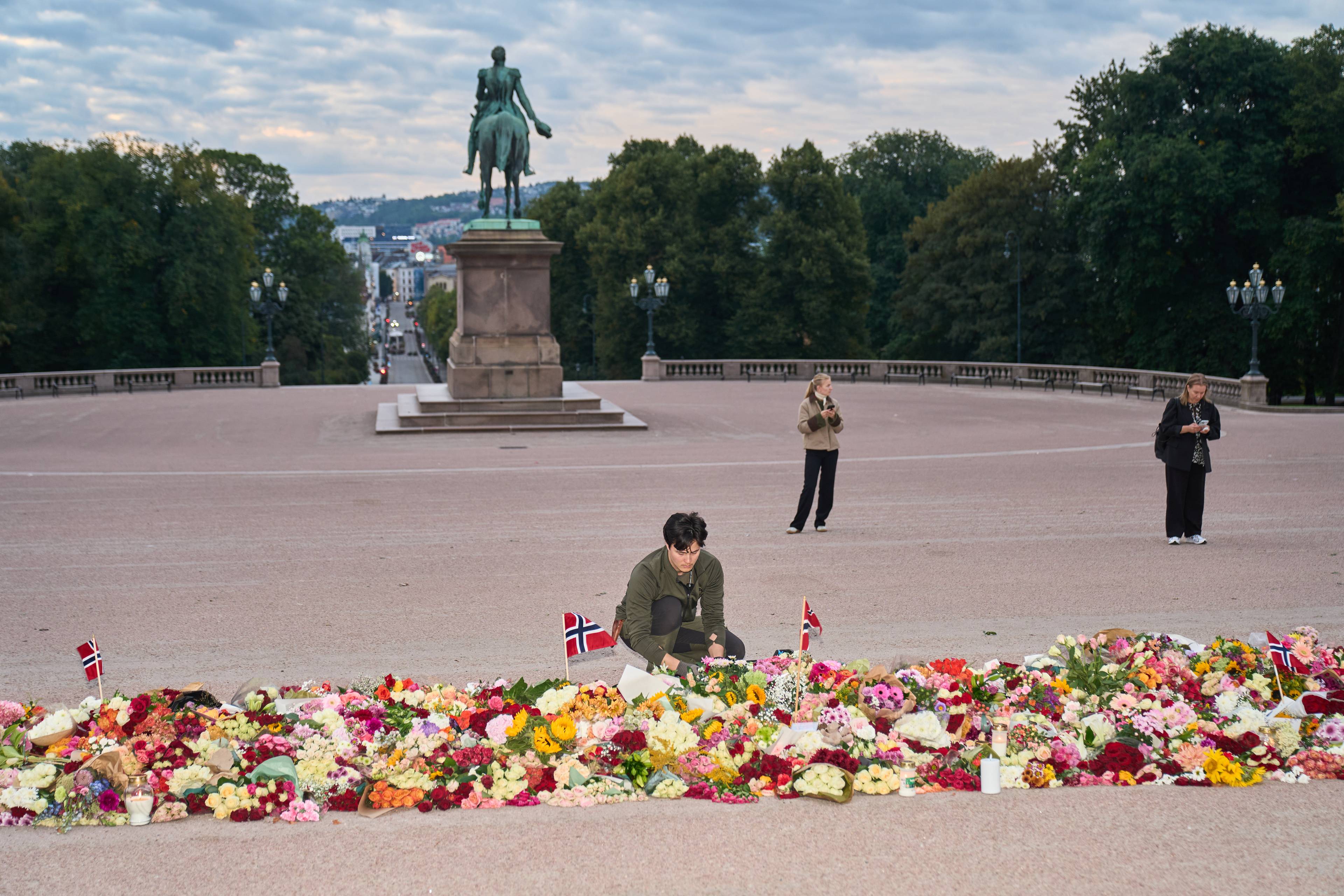 Person legger ned blomster foran Slottet i Oslo, omgitt av mange blomsterbuketter og norske flagg. (Bildebeskrivelsen er laget av en KI-tjeneste)