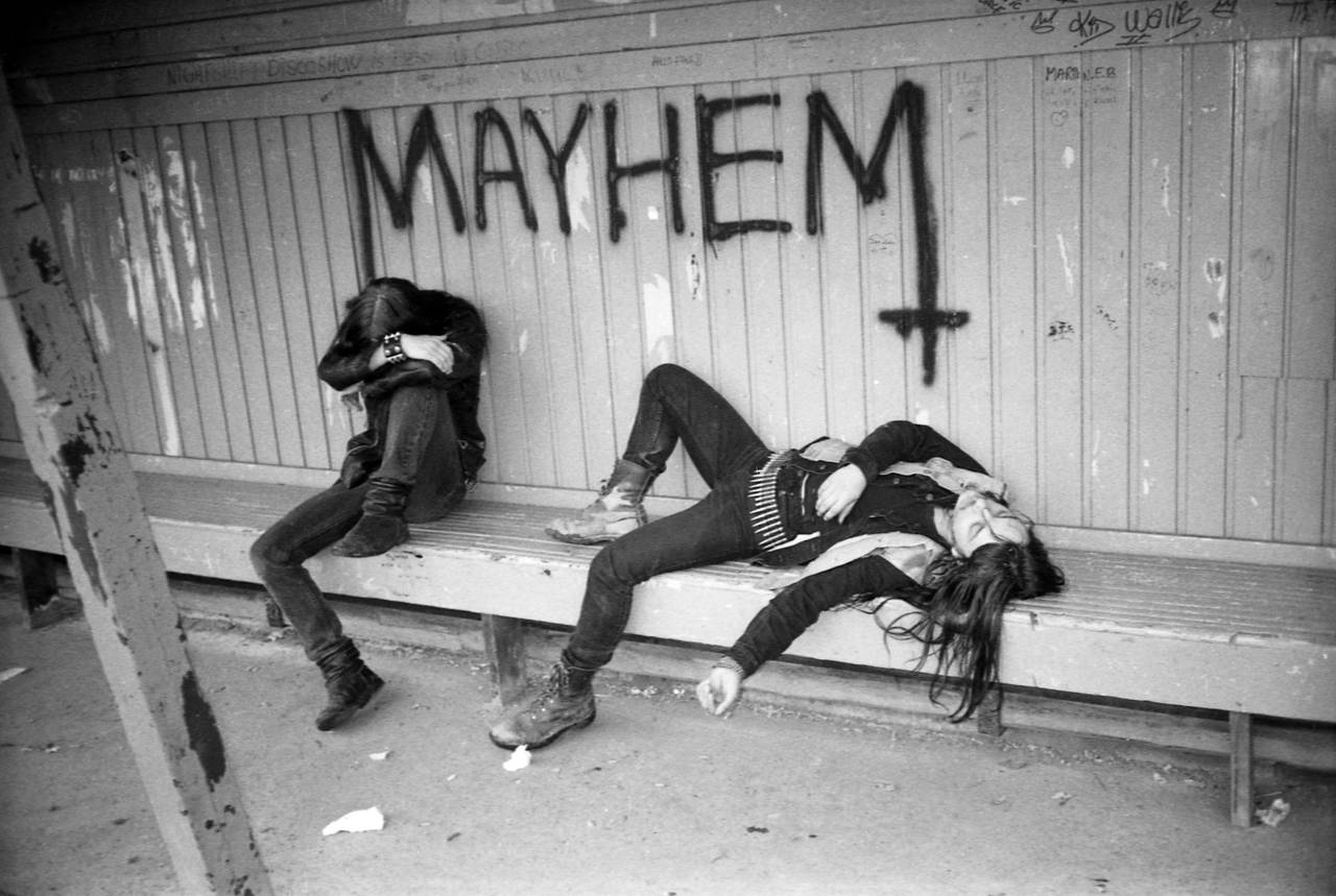 Øystein Aarseth sitter og Jørn Stubberud ligger på en benk på en togstasjon, hvor Mayhem-logoen er tagget på veggen bak dem.
