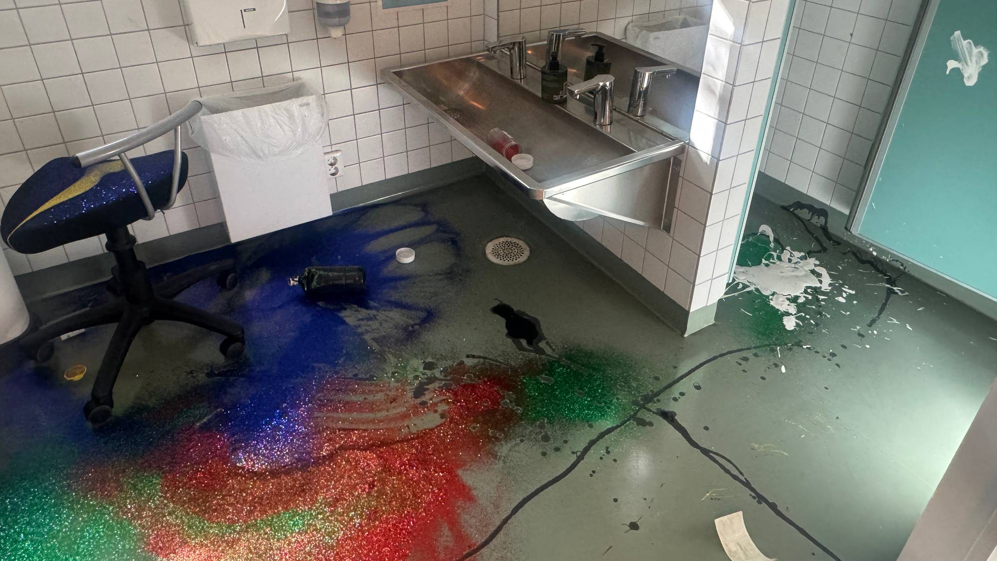 Søl av fargerik glittermaling på gulvet i et toalettrom med servant og kontorstol. (Bildebeskrivelsen er laget av en KI-tjeneste)